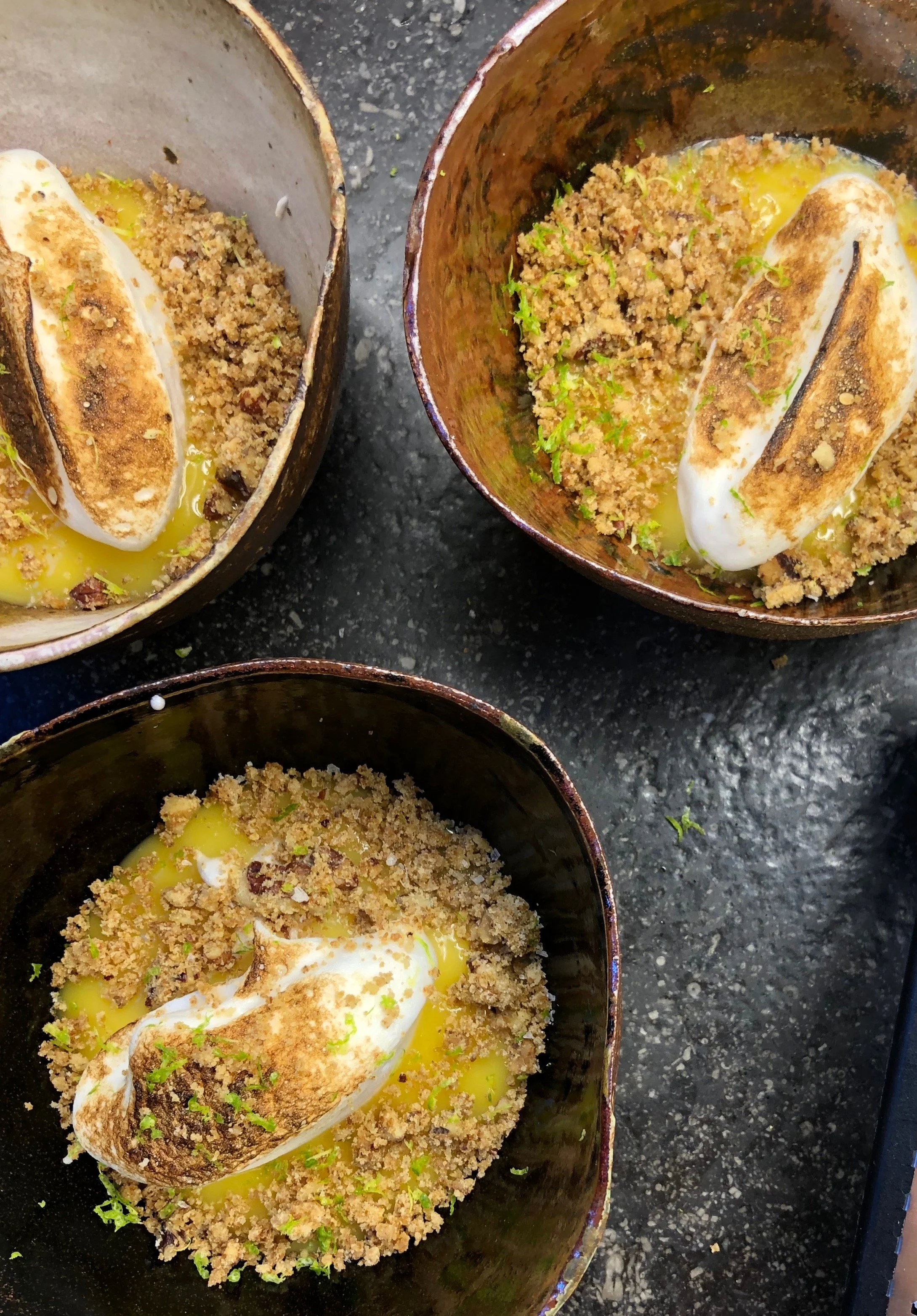 Three bowls of dessert topped with toasted marshmallow, crumbled graham crackers, and lime zest, served on a black surface.