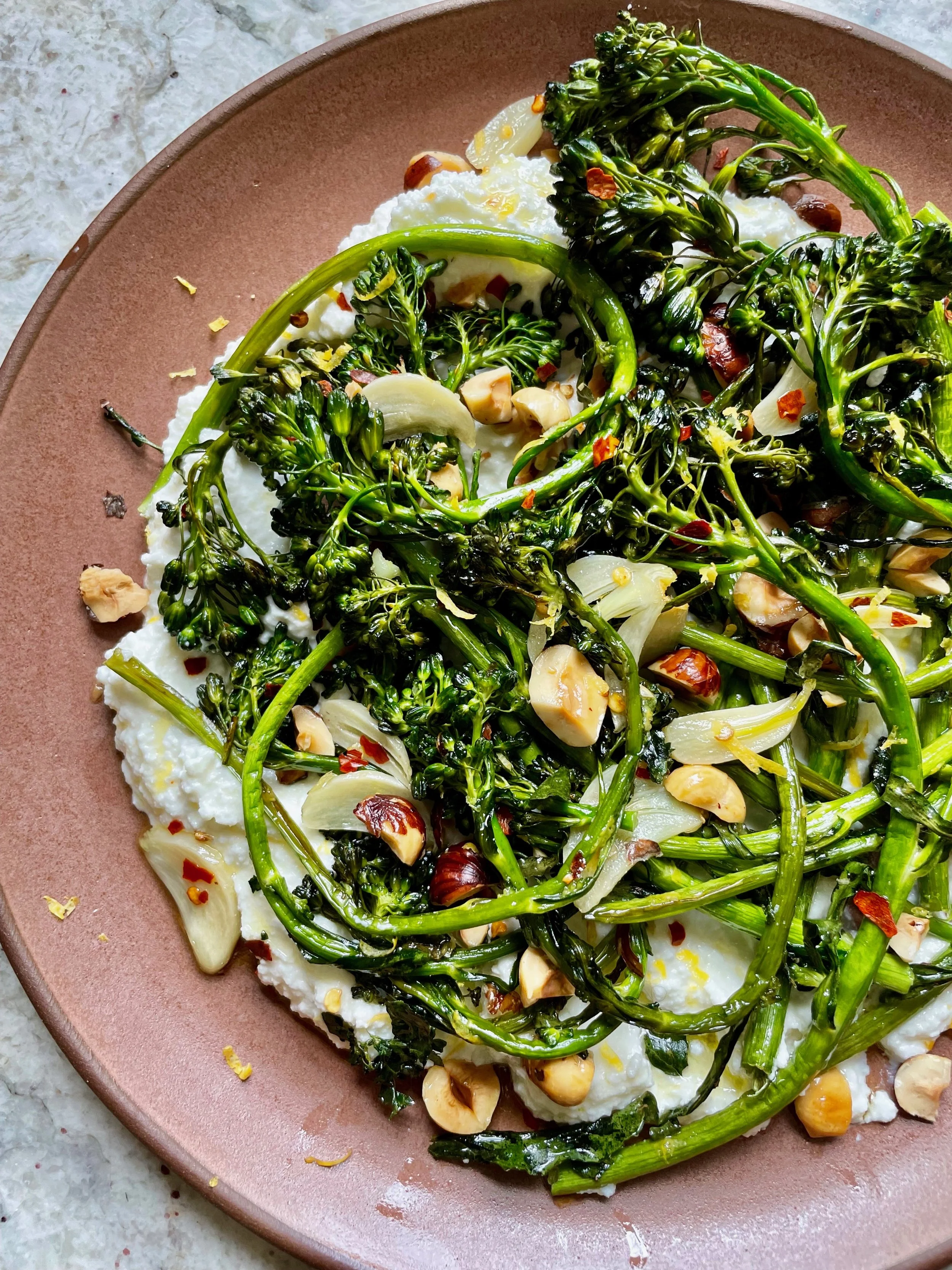 A plate of food containing roasted broccolini, sliced garlic, chopped nuts, and chili flakes served over white fluffy cheese or spread on a pink ceramic plate.