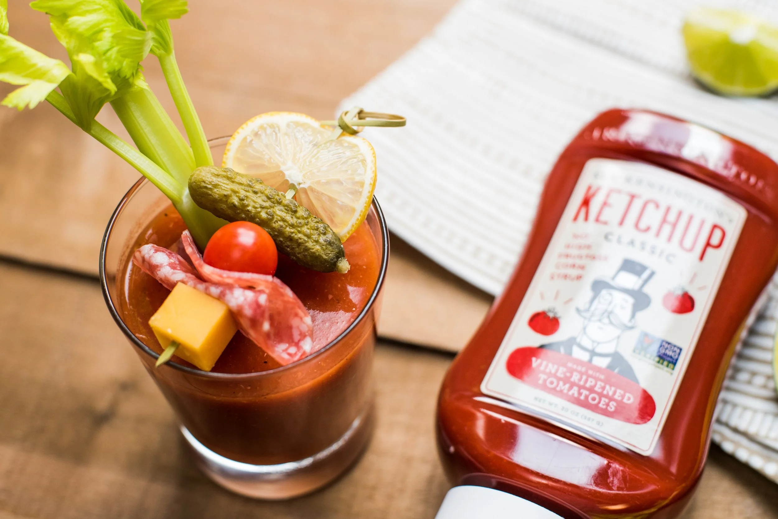 A glass of Bloody Mary cocktail garnished with lemon slice, cherry tomato, pickle, celery stalk, cheese cube, and a small piece of salami, next to a bottle of Hunt's ketchup on a wooden table.