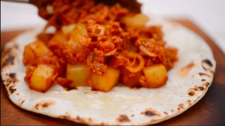 A flatbread topped with seasoned, cubed potatoes and a spicy sauce, placed on a wooden surface.