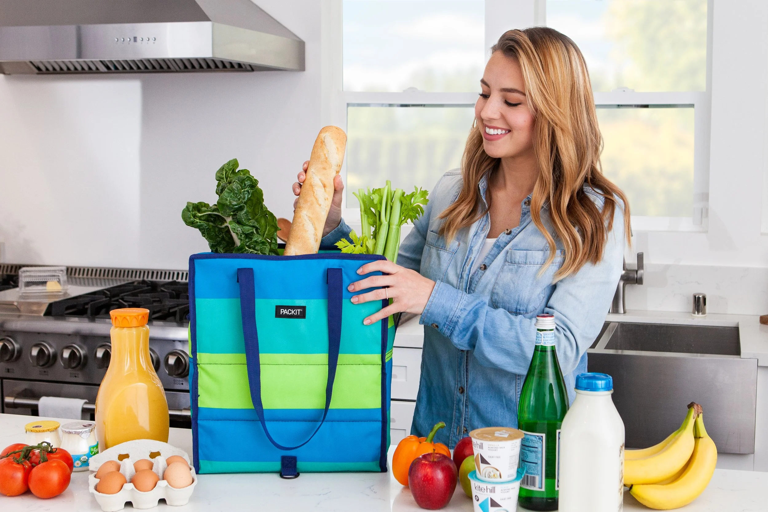 A woman smiling while placing a baguette into a reusable shopping bag full of green vegetables in a kitchen. The countertop has fresh tomatoes, eggs, bananas, a bottle of orange juice, yogurt, and other grocery items.
