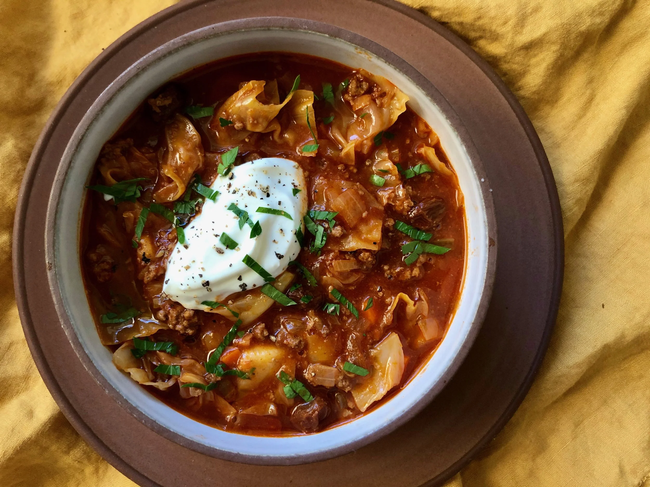 Bowl of beef and vegetable soup topped with sour cream, chopped chives, and black pepper, served on a brown-rimmed plate on a yellow cloth background.