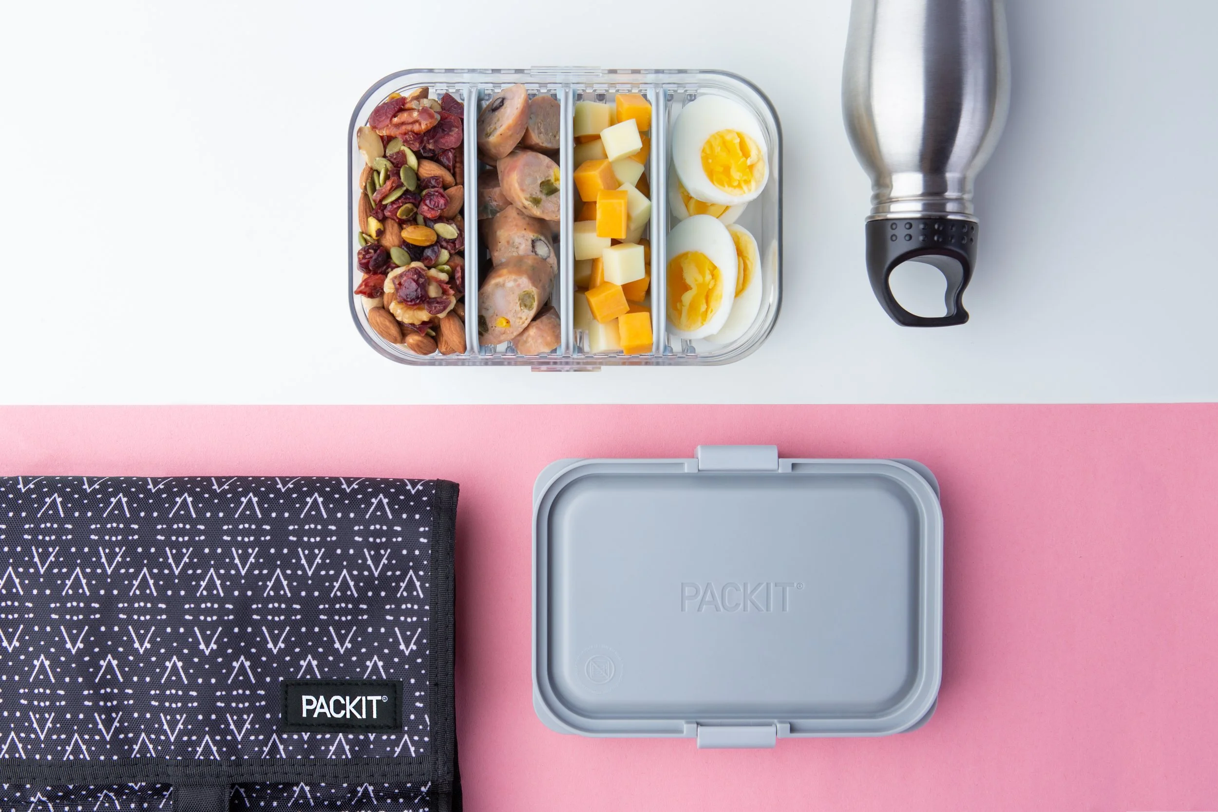 A packed lunch container with divided sections holding trail mix, sausages, cheese cubes, and boiled eggs, a metal water bottle, a black patterned lunch bag labeled 'PACKIT', and a gray lunchbox also labeled 'PACKIT', all placed on a pink and white s