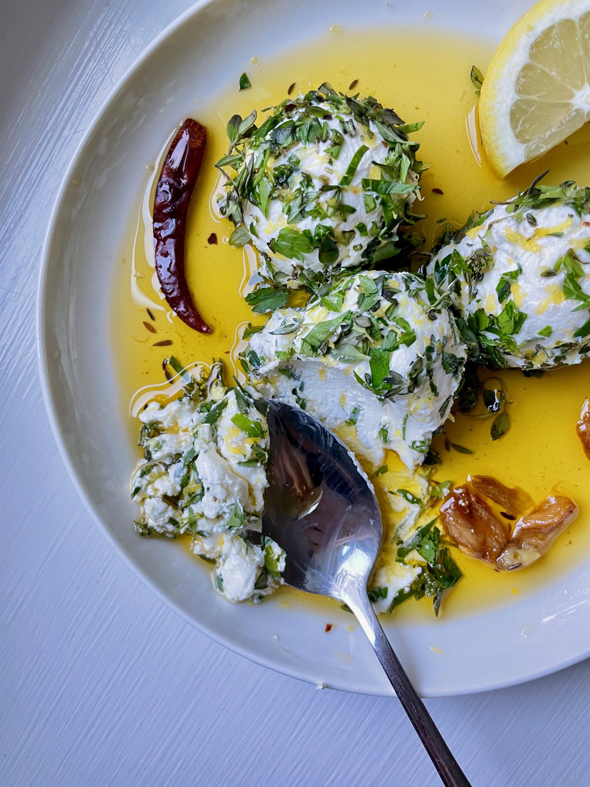 Close-up of a white plate with three stuffed eggs garnished with chopped herbs, drizzled with olive oil, and accompanied by a lemon wedge and a red chili pepper.