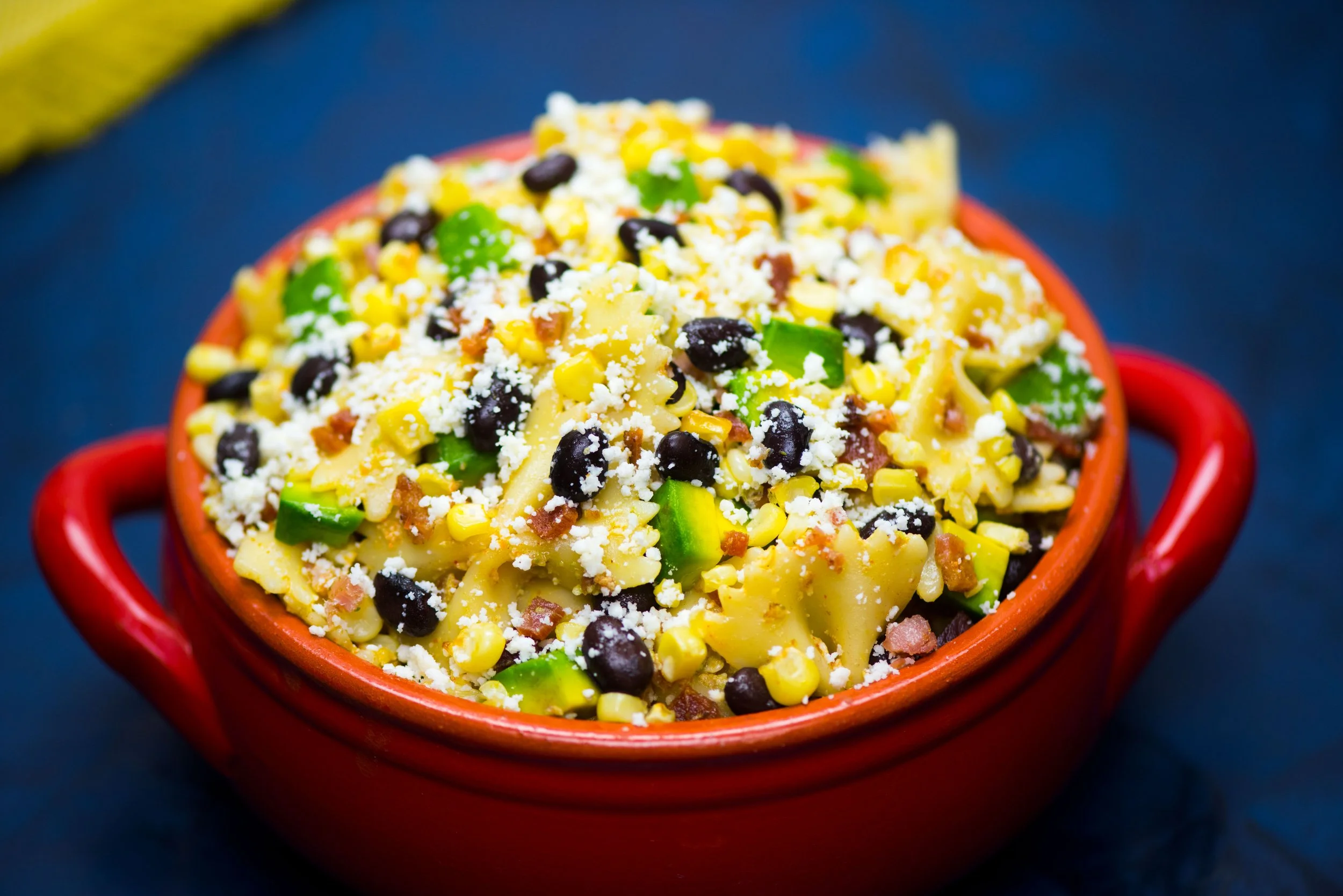 Close-up of a bowl of pasta salad with black beans, corn, chopped green peppers, shredded cheese, and small bits of bacon in a red bowl on a dark surface.