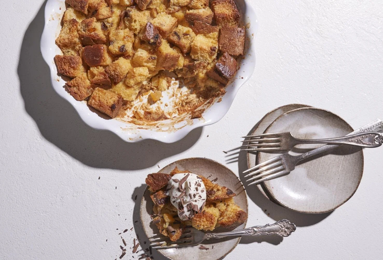 A slice of apple crumble with whipped cream and chocolate shavings on top, a larger bowl of apple crumble, and an empty plate with forks on a white surface.