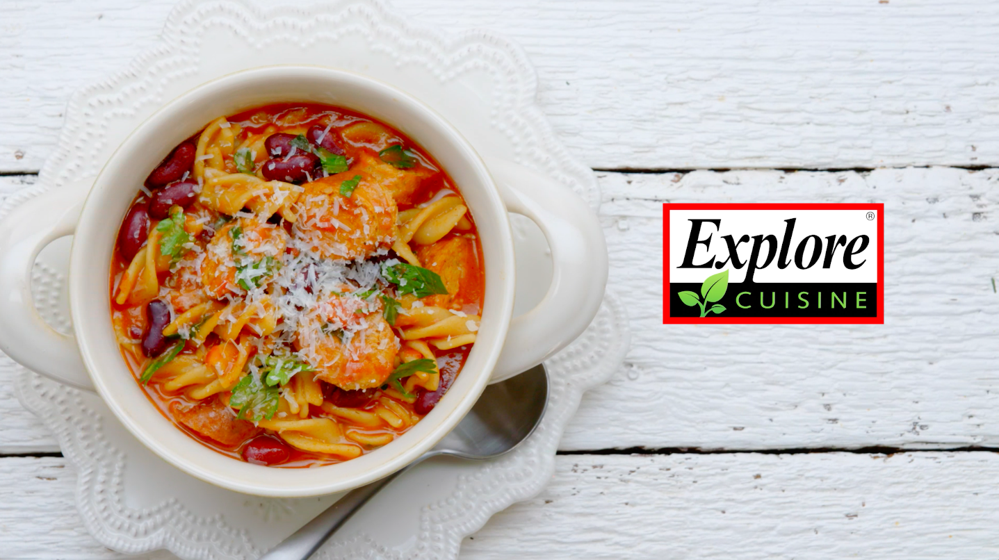 A bowl of chicken tortellini soup with beans, herbs, and grated cheese, on a white wooden surface with a white doily and a spoon, with a logo that says 'Explore Cuisine'