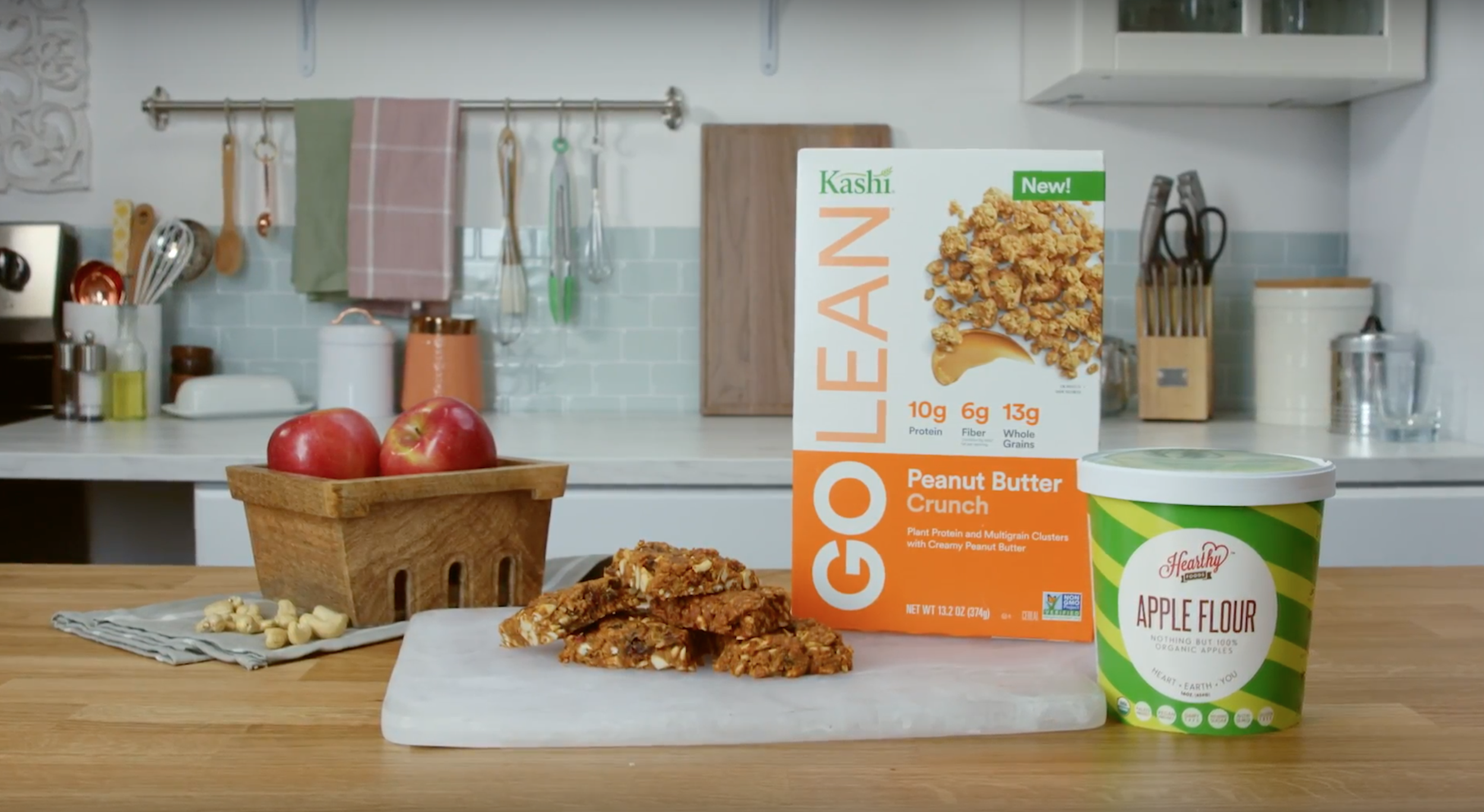 A box of Kashi GOLEAN Peanut Butter Crunch cereal, a container of Hearthy Apple Flour, a wooden bowl with three red apples, and a sliced granola bar on a cutting board on a kitchen countertop.