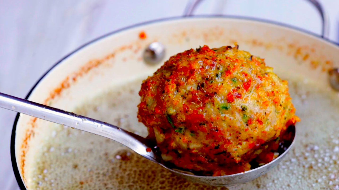 A close-up of a metal spoon holding a single meatball above a pot of creamy meatball soup or sauce.