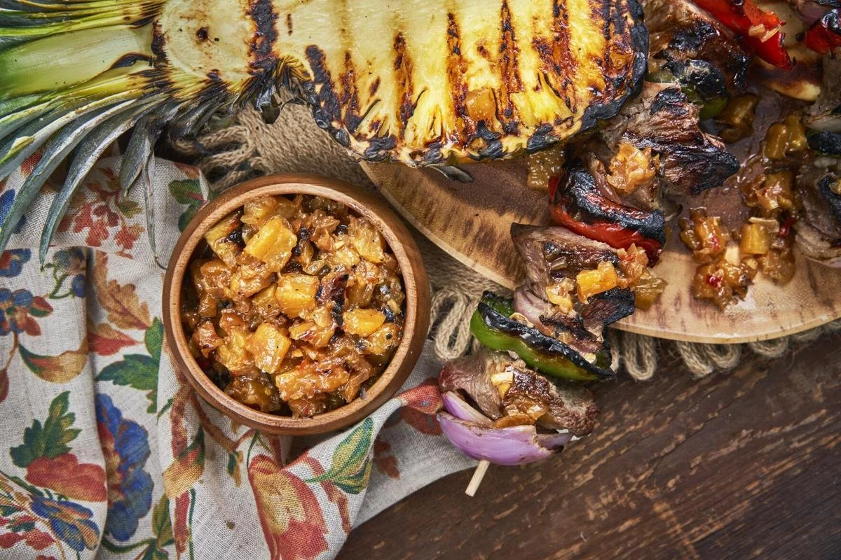 Grilled pineapple slice, chopped cooked vegetables, and beef kabobs on a wooden platter with a colorful cloth underneath.