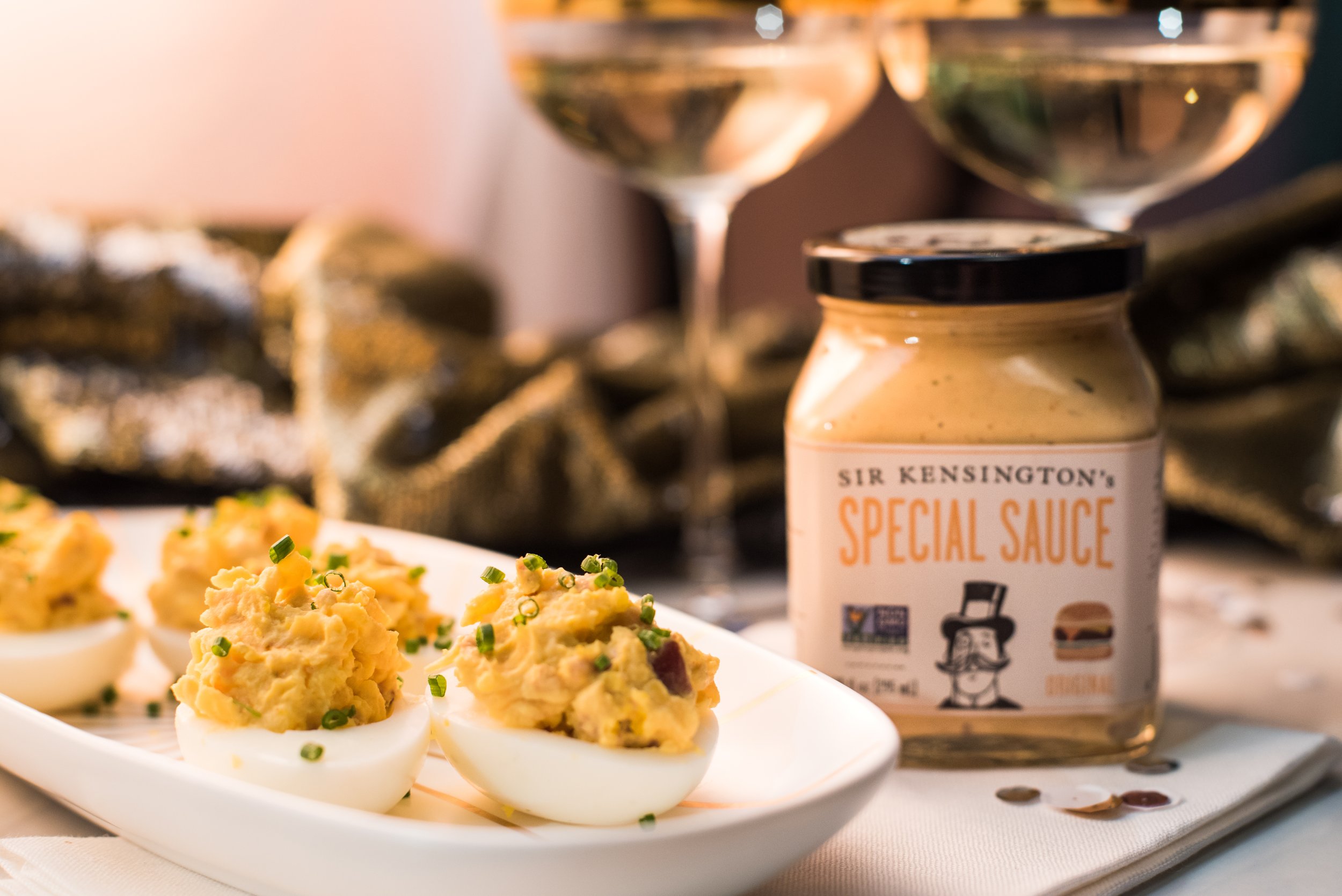Deviled eggs garnished with chopped chives, a jar of Sir Kensington's Special Sauce, and two glasses of white wine on a table setting.