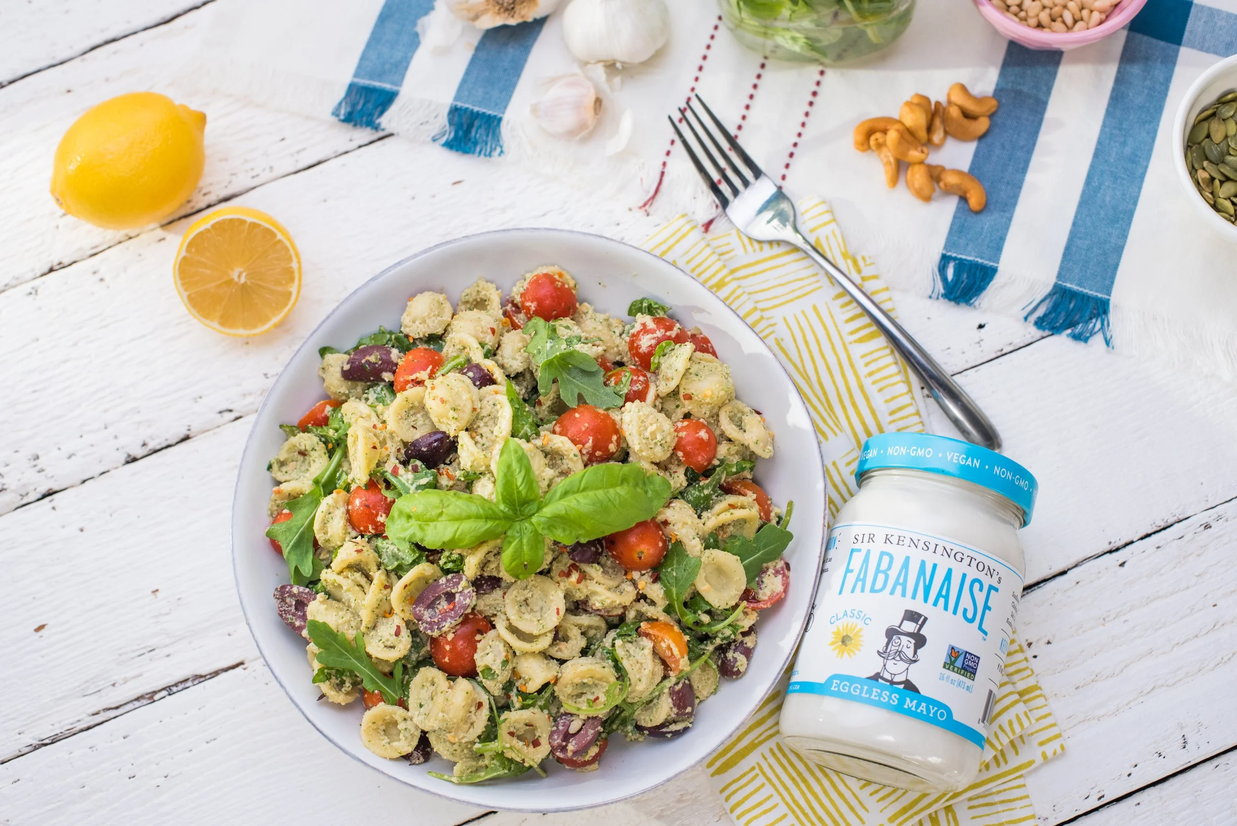 A bowl of pasta salad with cherry tomatoes, arugula, and basil, topped with a fork, and a jar of Sir Kensington's Tabasco vegan eggless mayo. Sliced lemon, garlic, cashews, and other ingredients are on a white wooden table with a blue and yellow stri