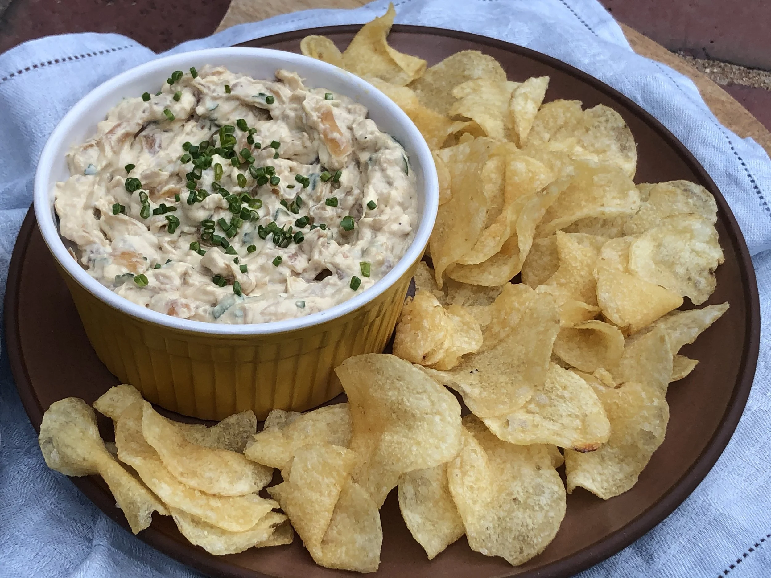 A yellow bowl of potato salad garnished with chopped chives, served with a side of potato chips on a brown tray.