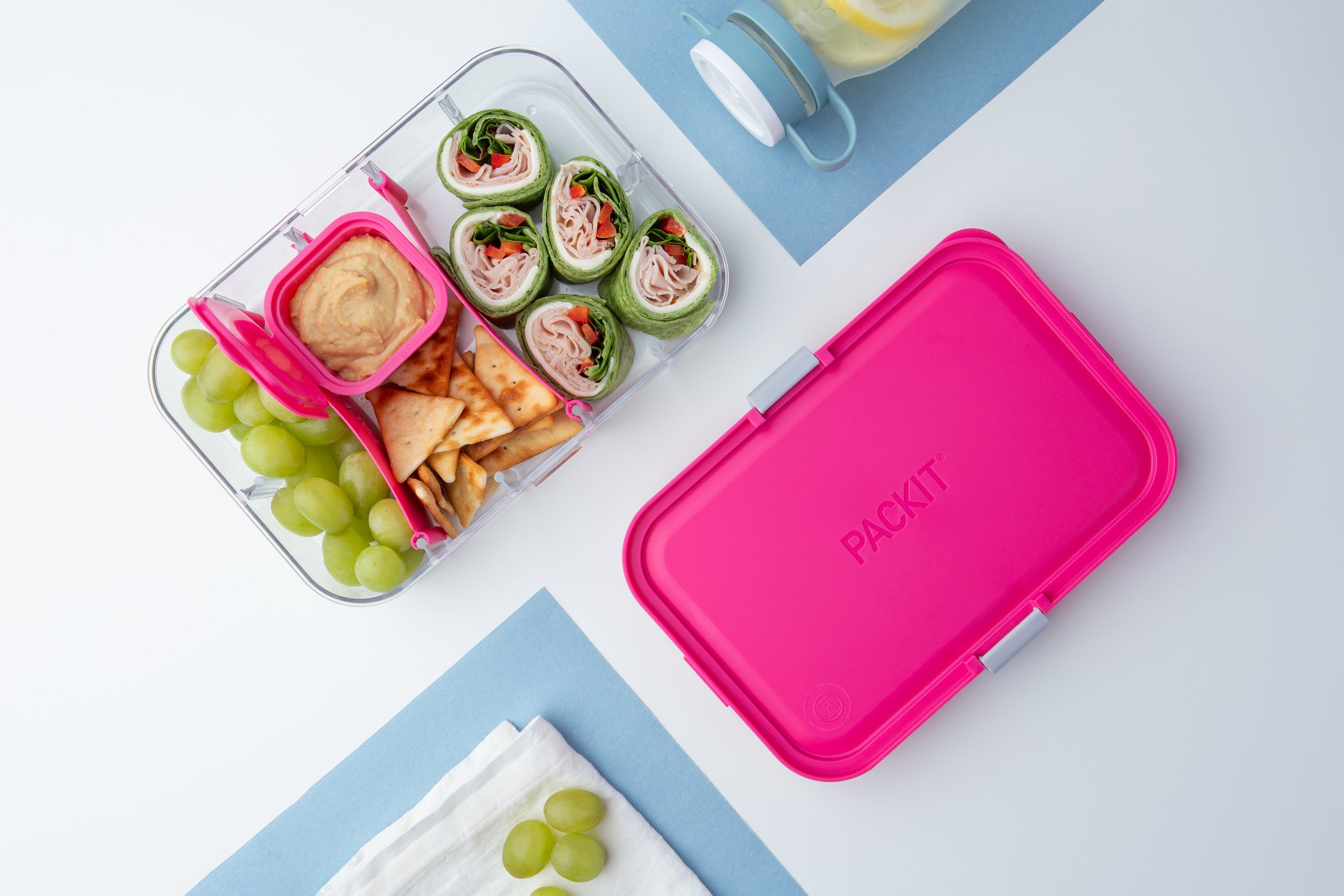 A lunchbox containing grapes, hummus with crackers, and cucumber roll-ups with turkey and vegetables, next to a pink PACKIT lunchbox on a white surface with a blue placemat and a beverage bottle.