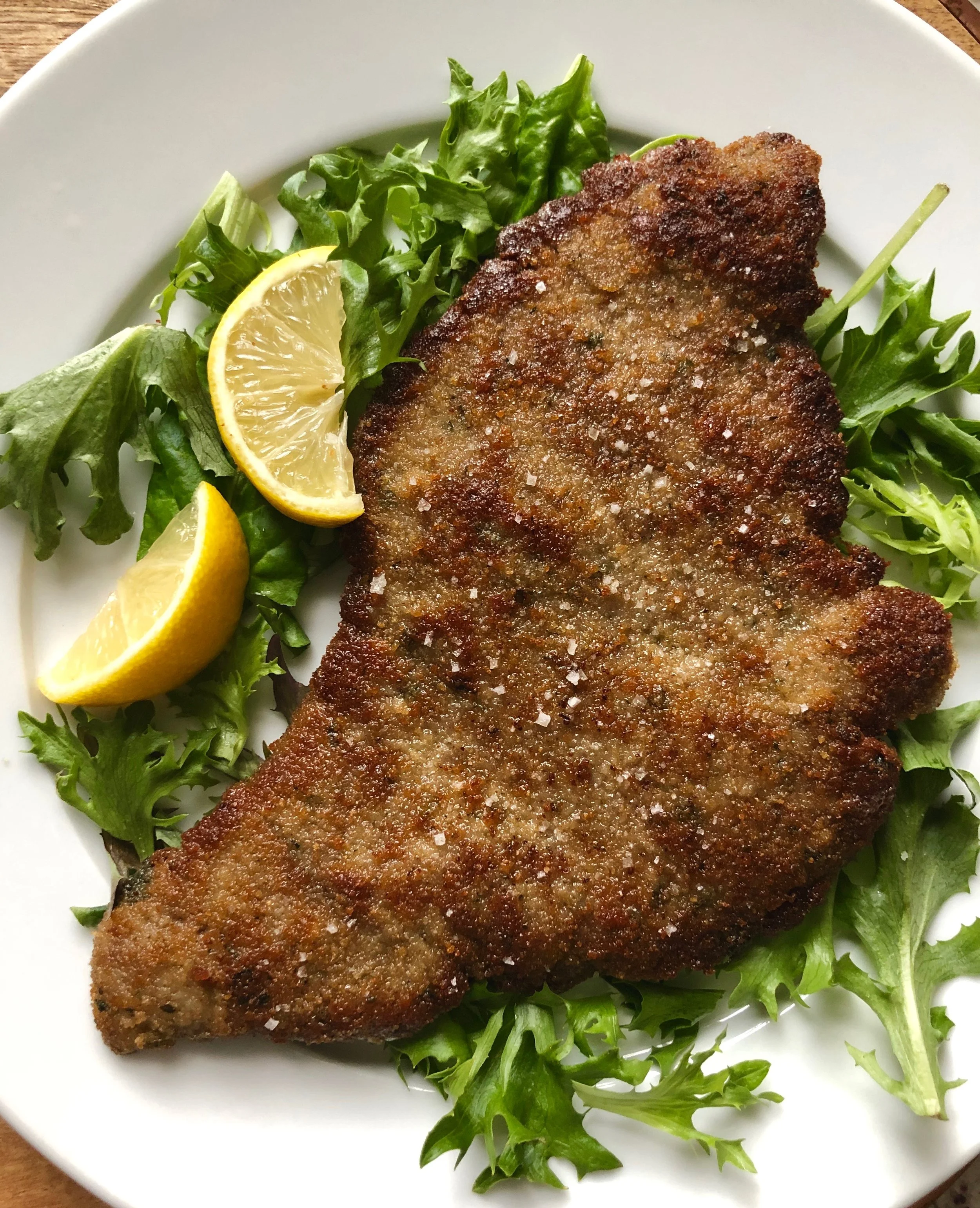 Breaded and fried fish fillet garnished with coarse salt, served on a bed of mixed greens with lemon wedges on a white plate.