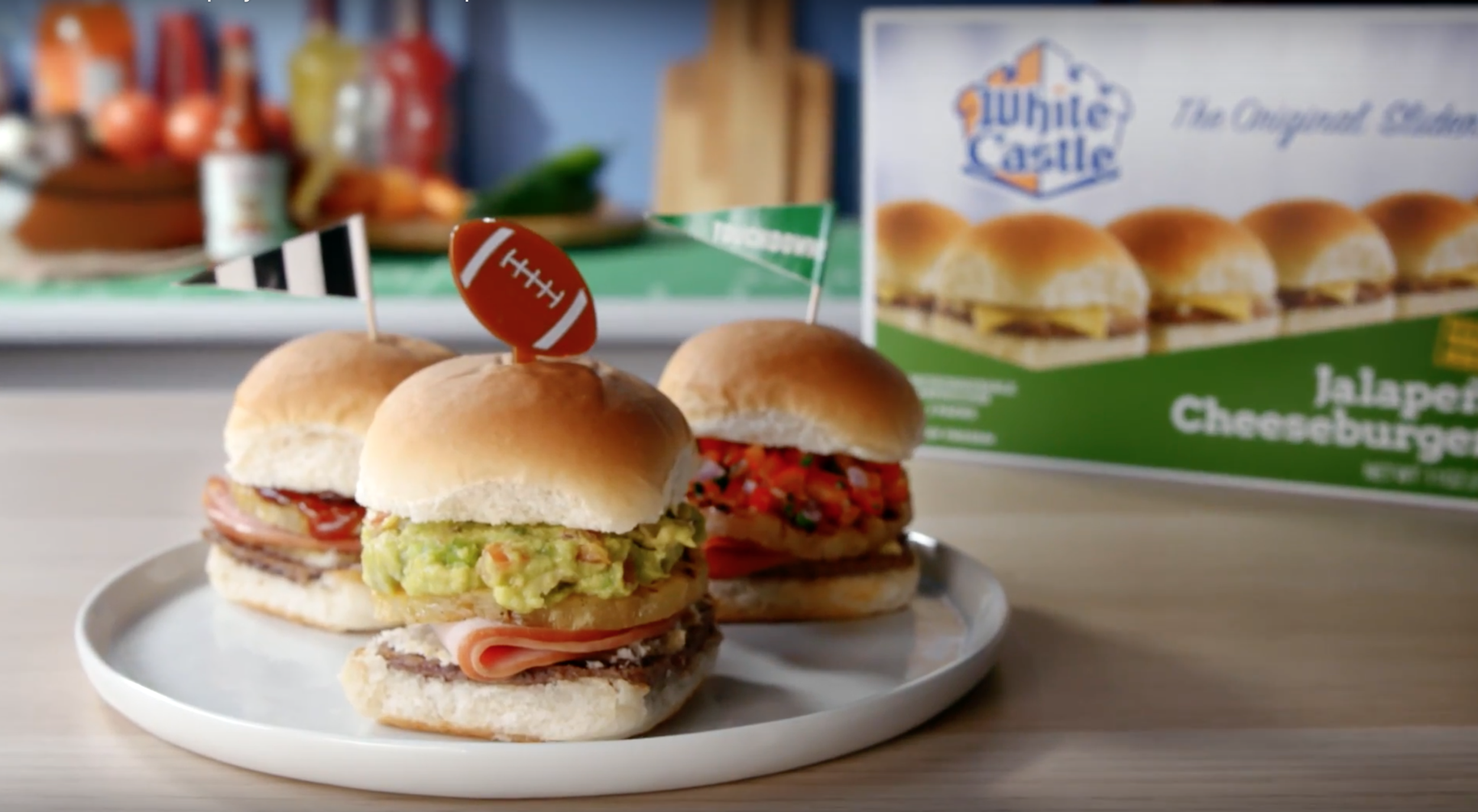 Three mini sandwiches with various fillings on a white plate in front of a green box of hamburger buns, with a background of condiments and other food items.