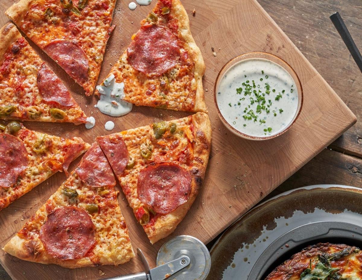 A wooden table with a pizza that has slices of pepperoni and green peppers, a pizza cutter, a bowl of creamy white dipping sauce garnished with chopped herbs, and parts of a glass bowl with another dish in the bottom right corner.