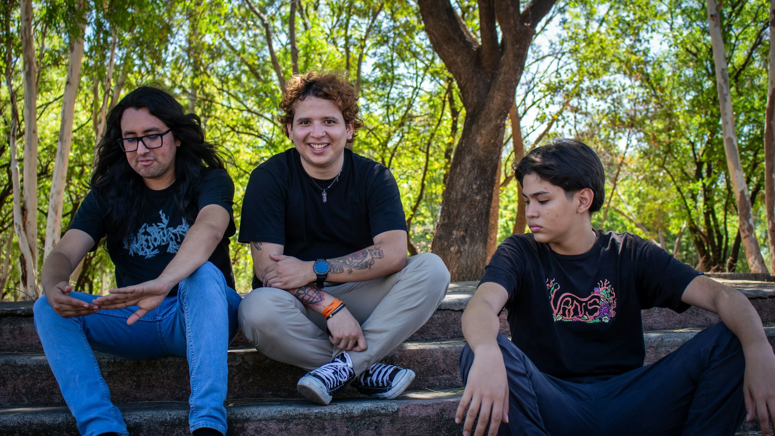 Three young men sitting on outdoor steps, surrounded by trees, one looking at his phone, one smiling, and one looking down, all dressed casually.