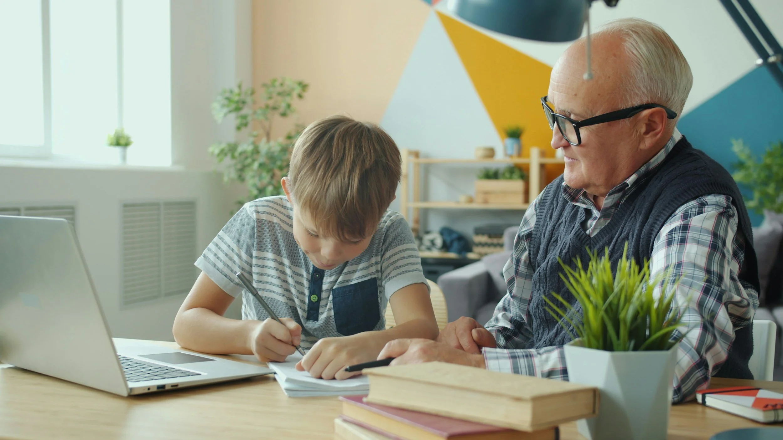 A young boy and an elderly man sit at a wooden table in a bright room, engaging in a learning activity. The boy is focused on writing in a notebook with a pen, while the elderly man looks on, observing. There are books, a potted plant, a laptop, and a notebook on the table, with a colorful geometric wall and a window with natural light in the background.