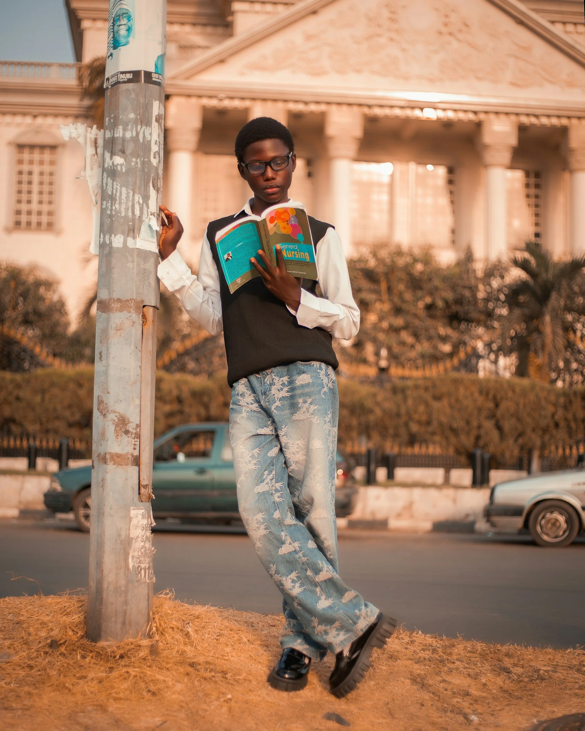 A young person with short hair, wearing glasses, a white shirt, black vest, and patterned jeans, leans against a pole while reading a book titled 'General Nursing' on a city street.