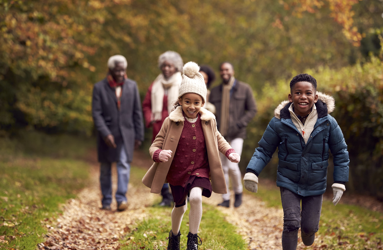 Group of children and elderly people walking and running on a leaf-covered trail in a park during autumn, surrounded by colorful fall trees.