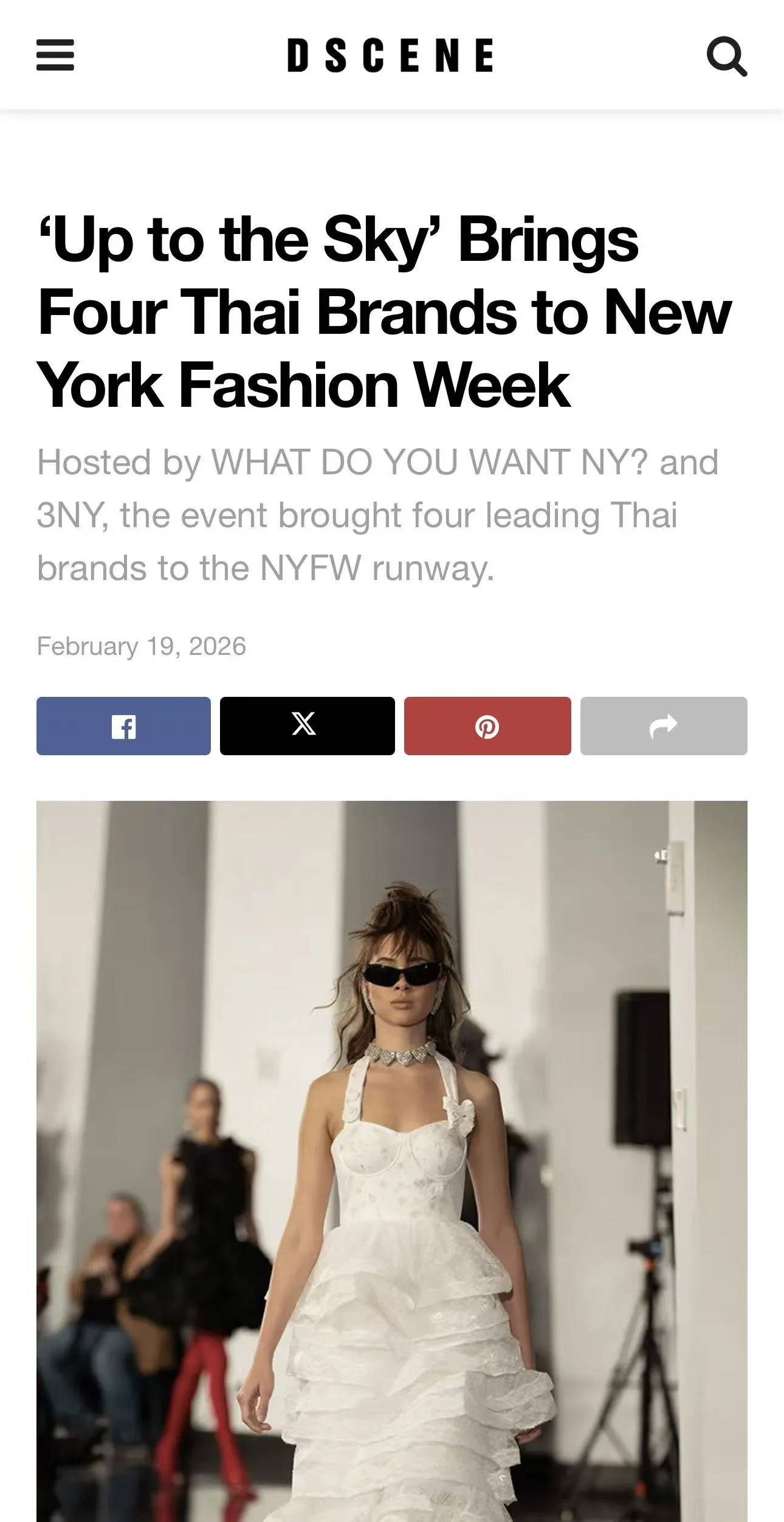 A model walking on a runway wearing a white ruffled dress and black sunglasses, with some audience members visible in the background.