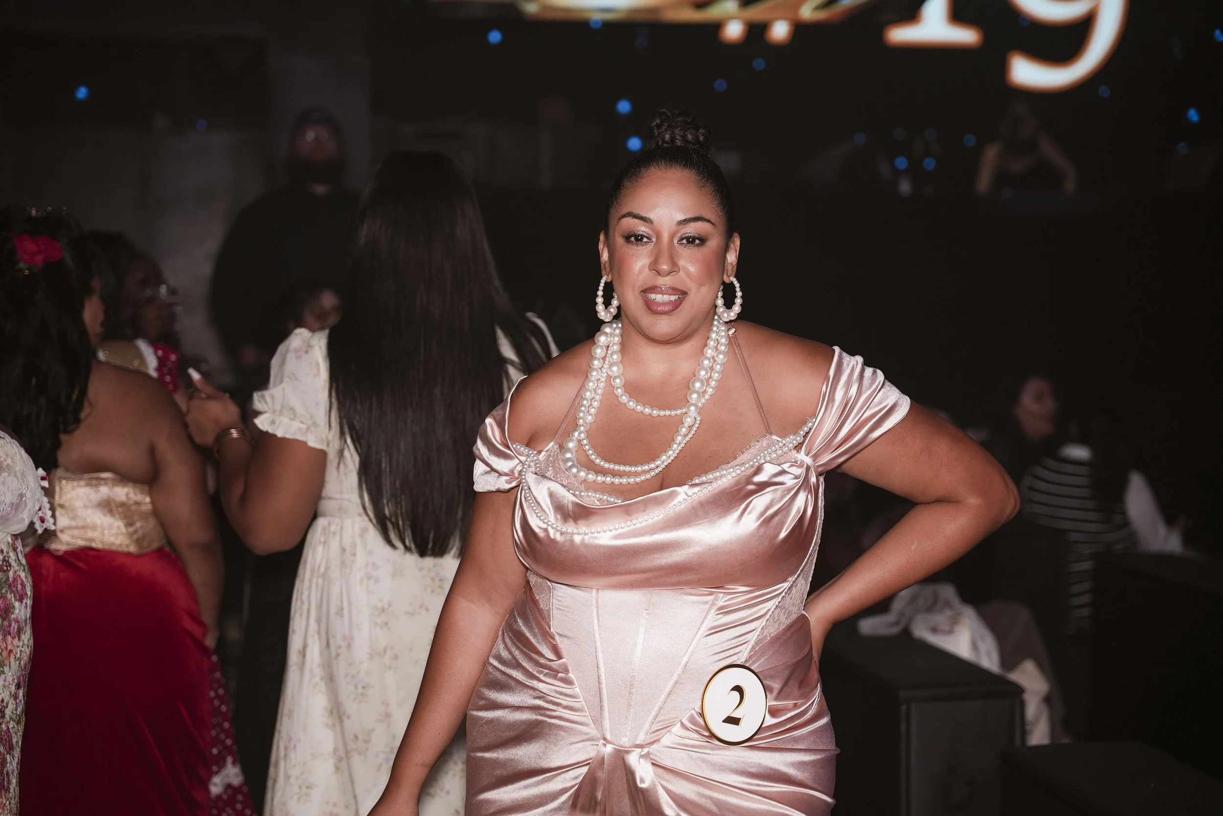 A model wearing a pink satin Lacemade dress, posting for Miss Curvy Pageant at the club Nebula, a NYFW runway show from the dating app WooPlus by event production agency ANG Concept.