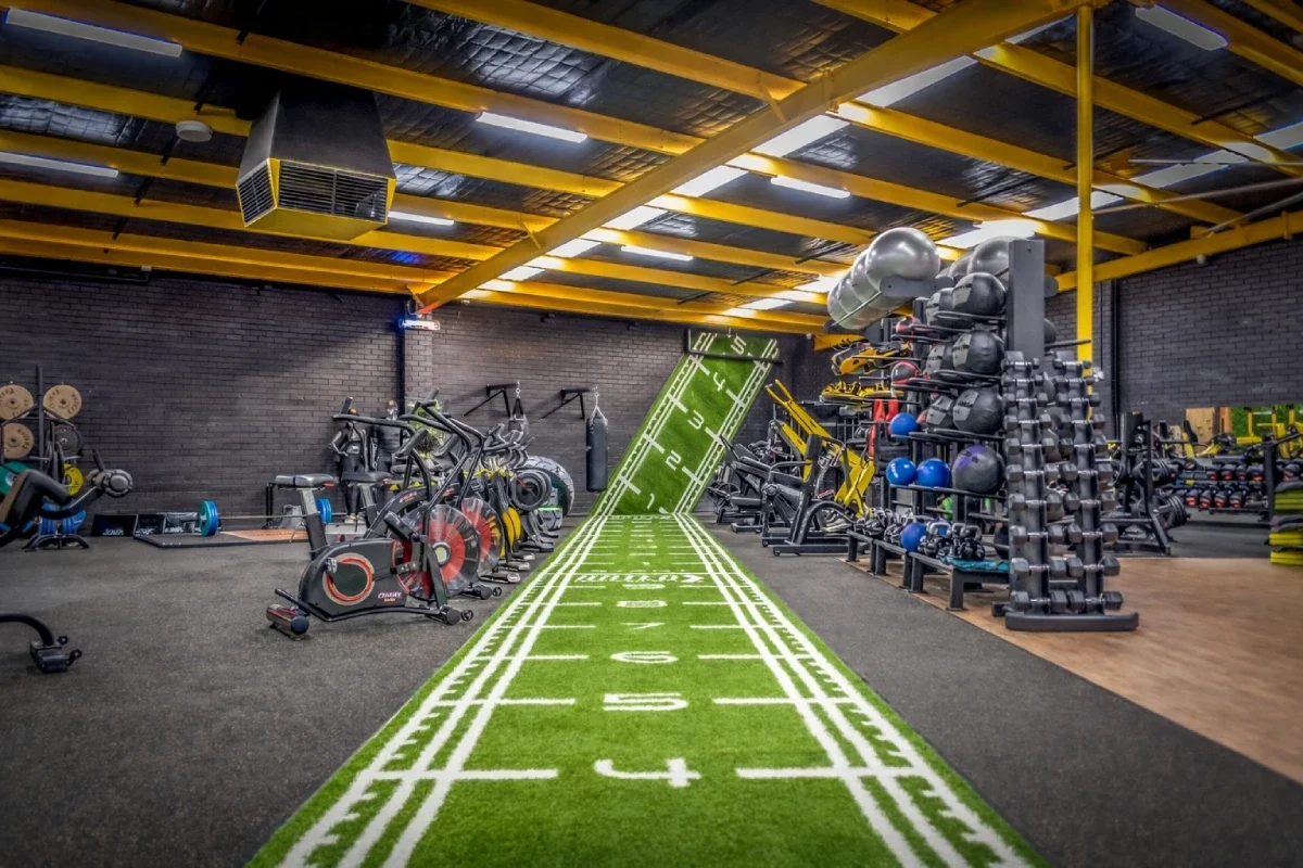 Indoor gym with artificial running track designed like a football field with yard markings, surrounded by various exercise equipment including stationary bikes, weight racks, medicine balls, and kettlebells, with yellow beams on the ceiling.