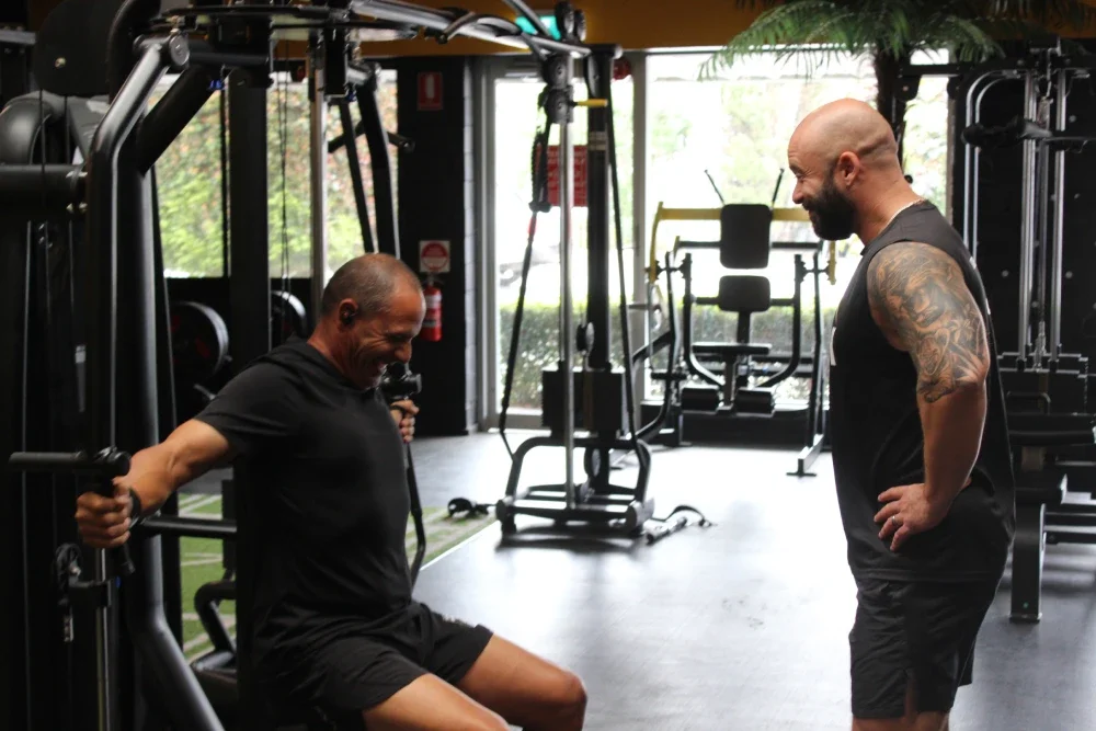 A man using a gym machine, working out, while another man trains and guides him in a gym with various workout equipment.