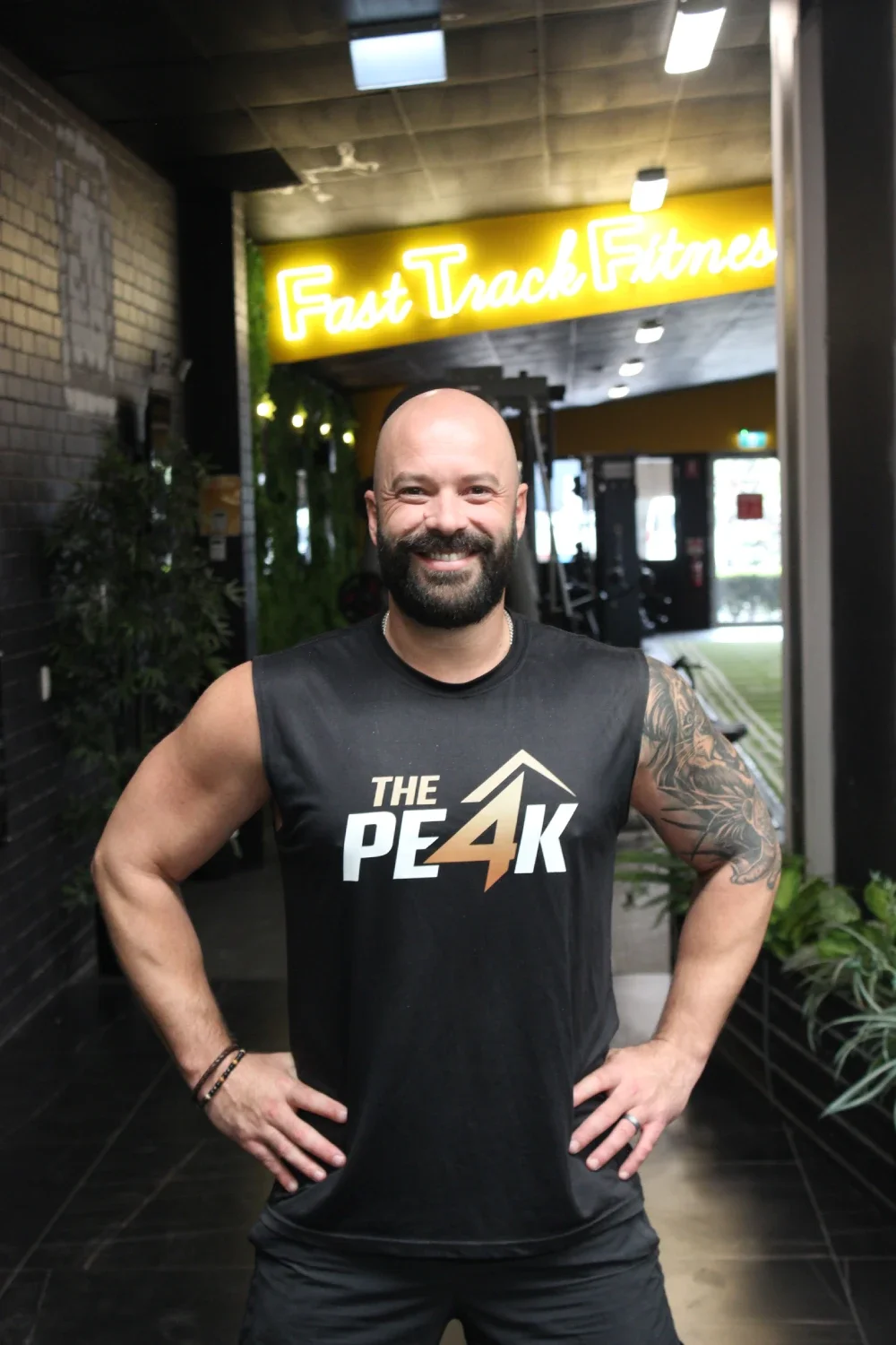 Gym Founder Adam smiling, wearing a black sleeveless shirt that says 'The Peak,' standing in a gym.
