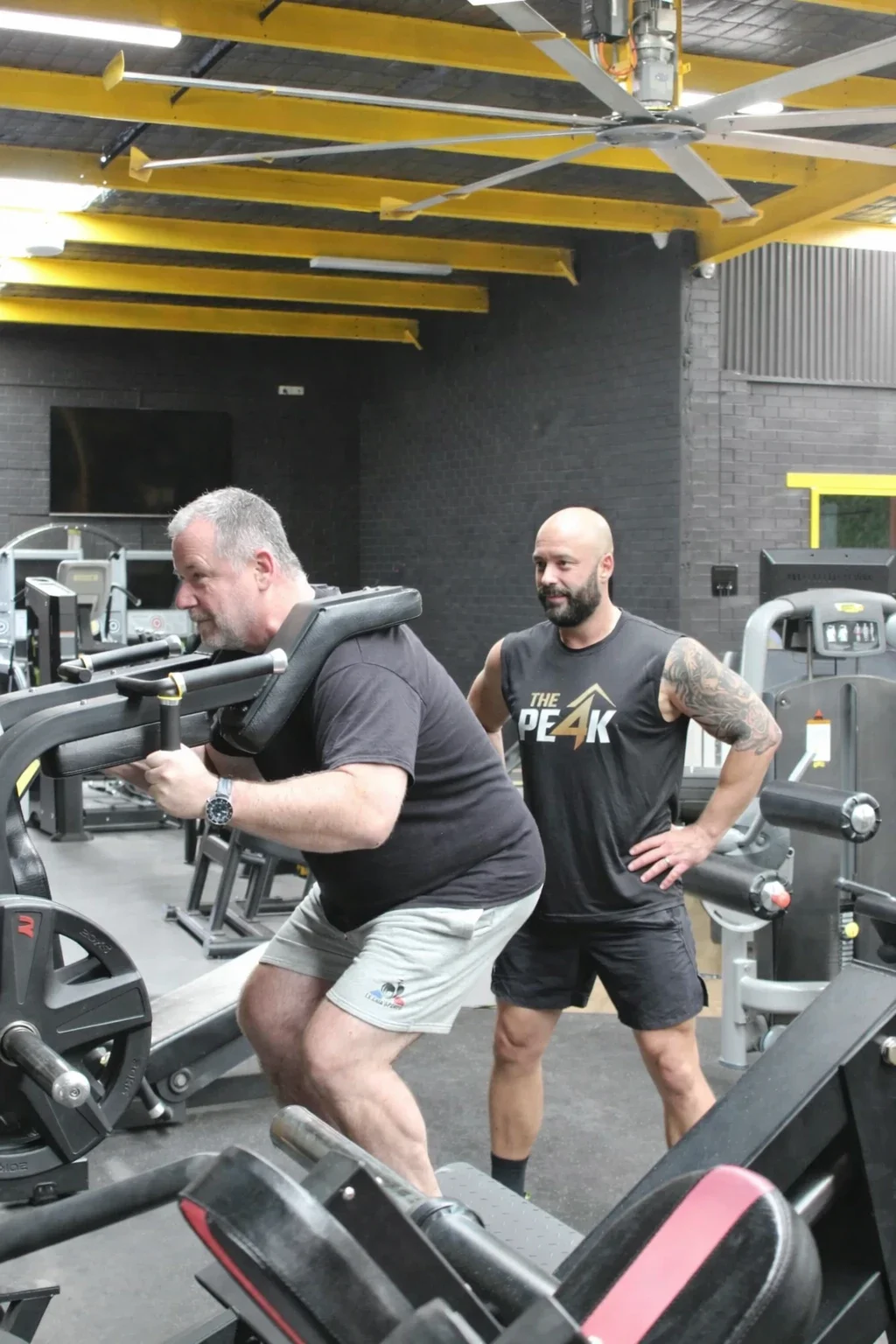 A man is lifting a weight with a machine in a gym, while another man observes and encourages him.