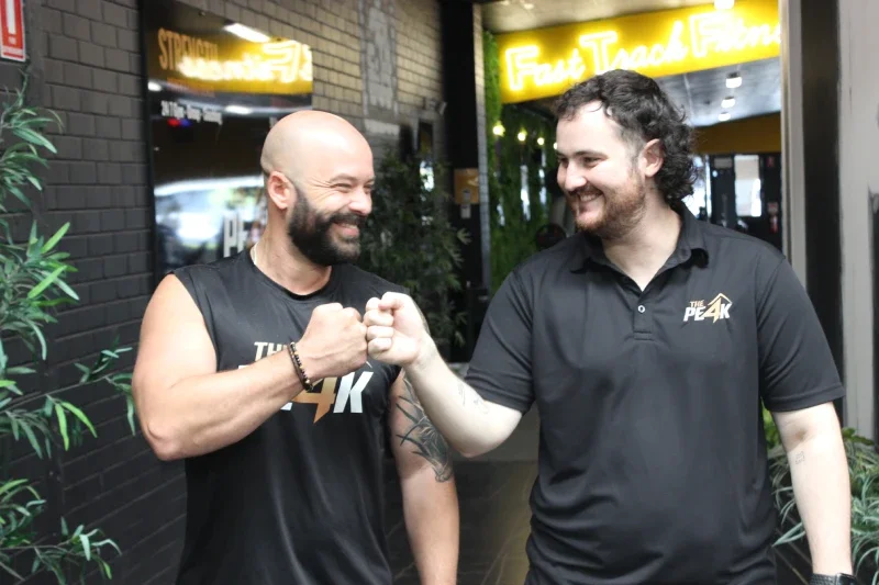 Two men are doing a fist bump and smiling. They are both gym staff wearing 'The Peak' shirts.