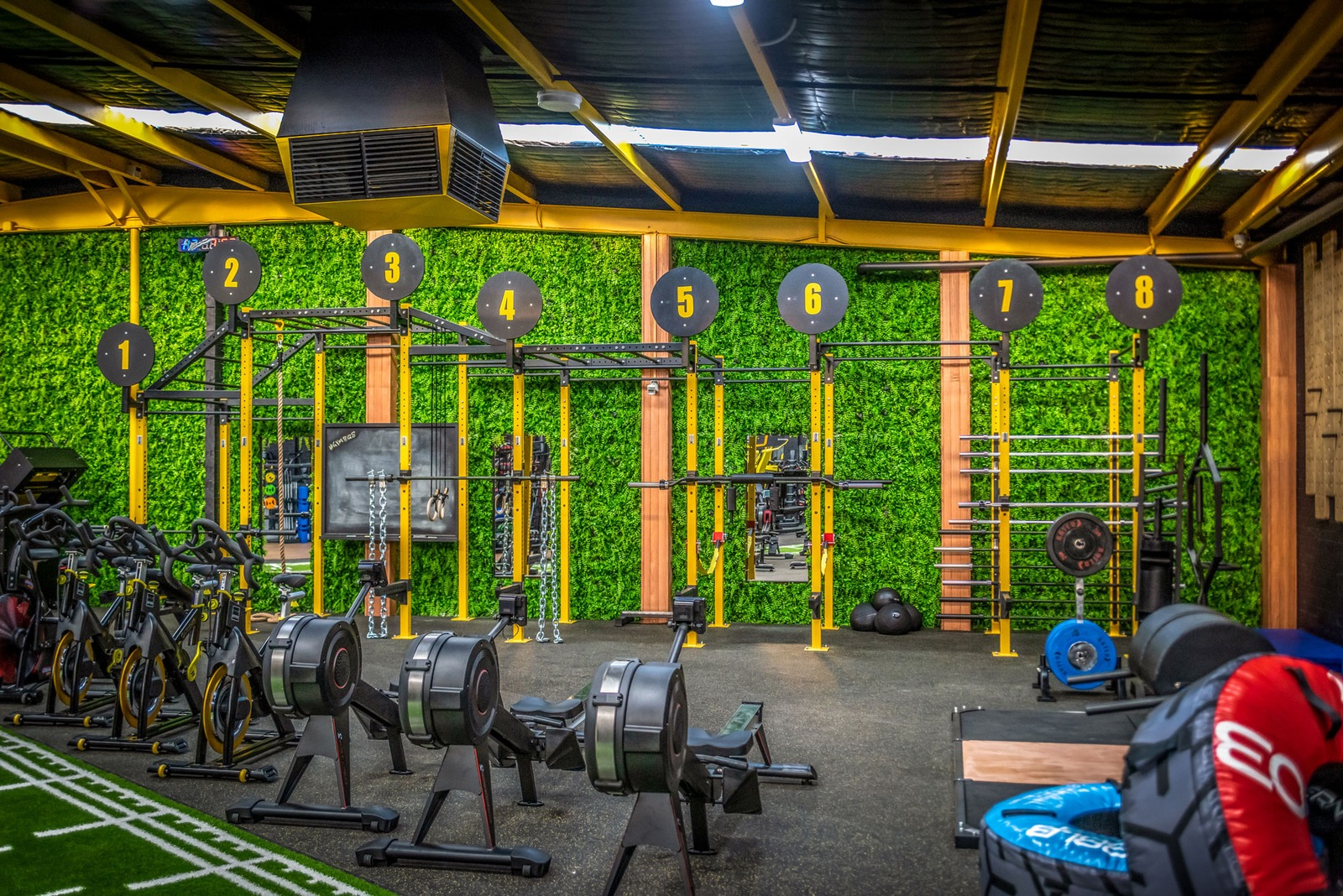 Fitness gym with various workout equipment, including rowing machines, weights, and pull-up bars, in front of a green wall with numbered circular targets from 1 to 8 on the upper part.