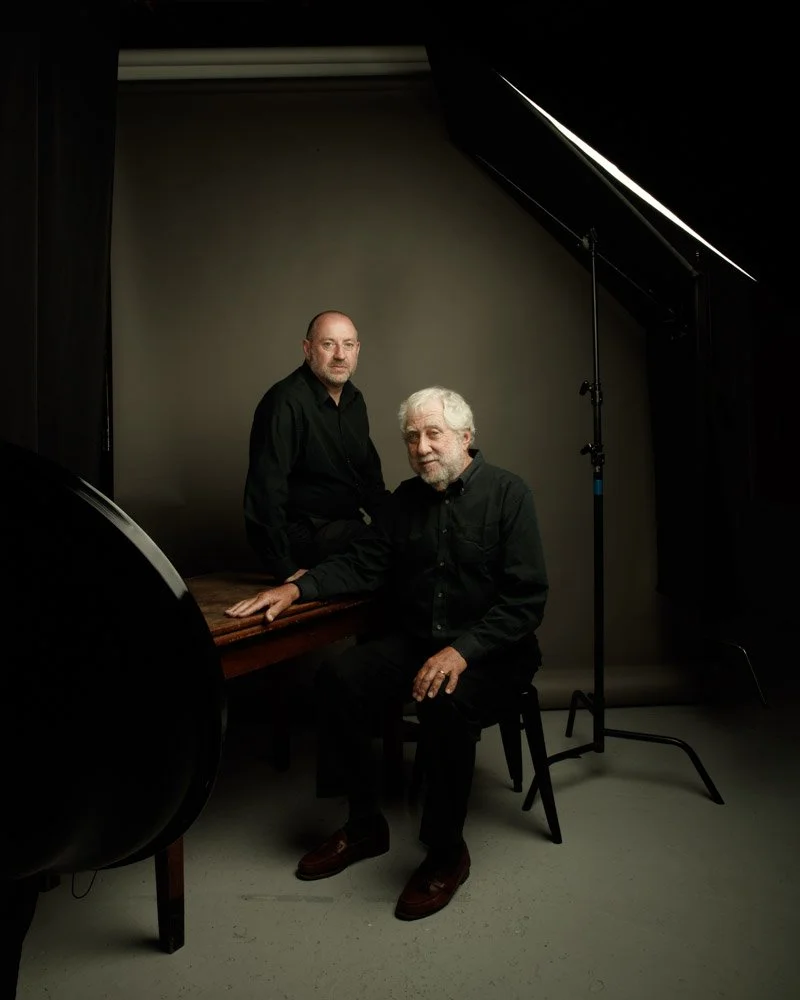 Two men in black shirts posing in a photography studio with professional lighting and a gray backdrop.
