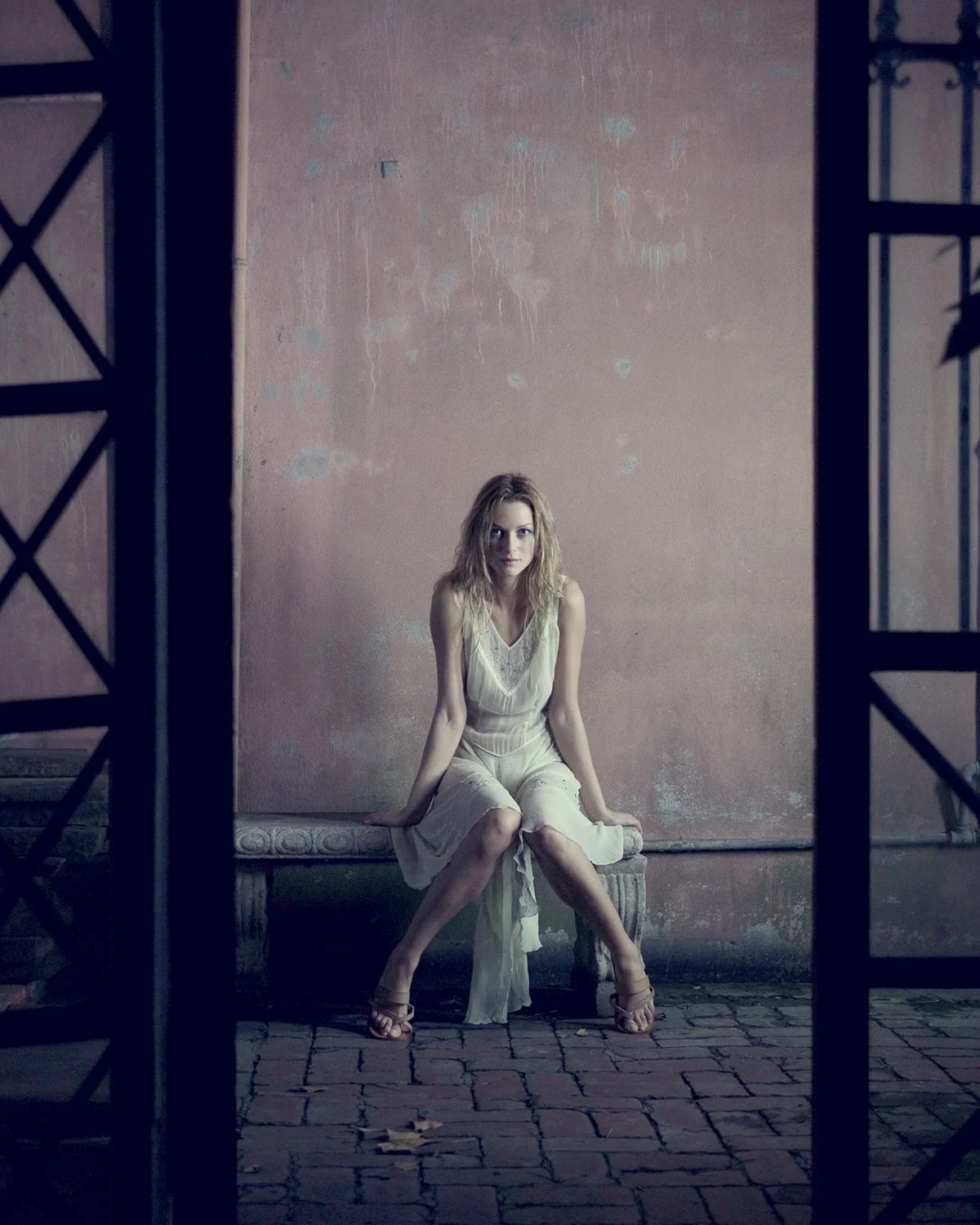 A woman sitting on a stone bench in front of a worn pink wall, framed by a metal gate.
