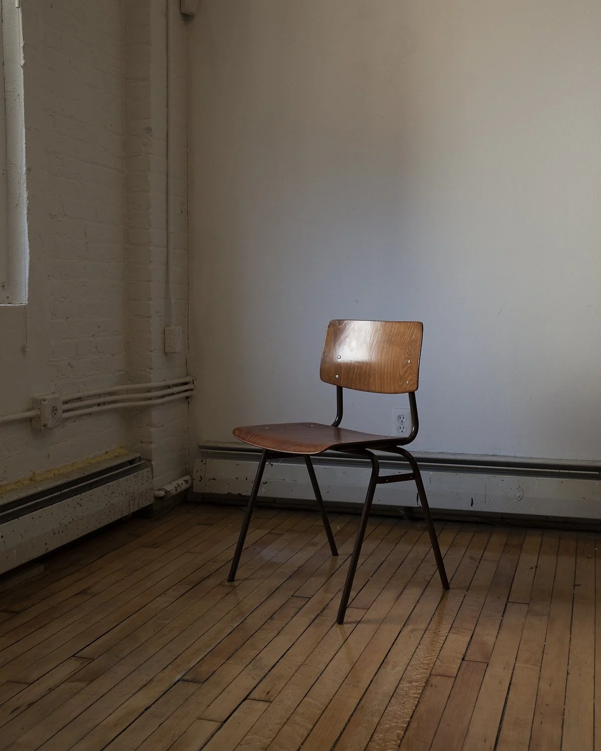 An empty wooden chair in a room with hardwood floors and exposed piping along the wall.