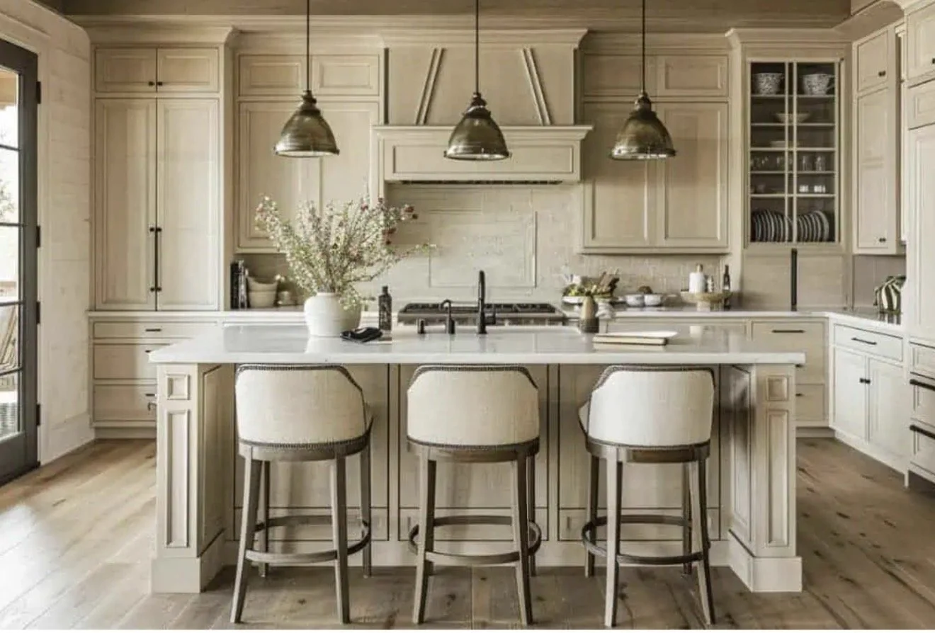 A modern farmhouse kitchen with a central island, three beige upholstered bar stools, and light wood cabinetry. There are three pendant lights hanging over the island, a stainless steel sink with a black faucet, and a vase of flowers on the countertop. To the left, a glass door allows natural light into the space.