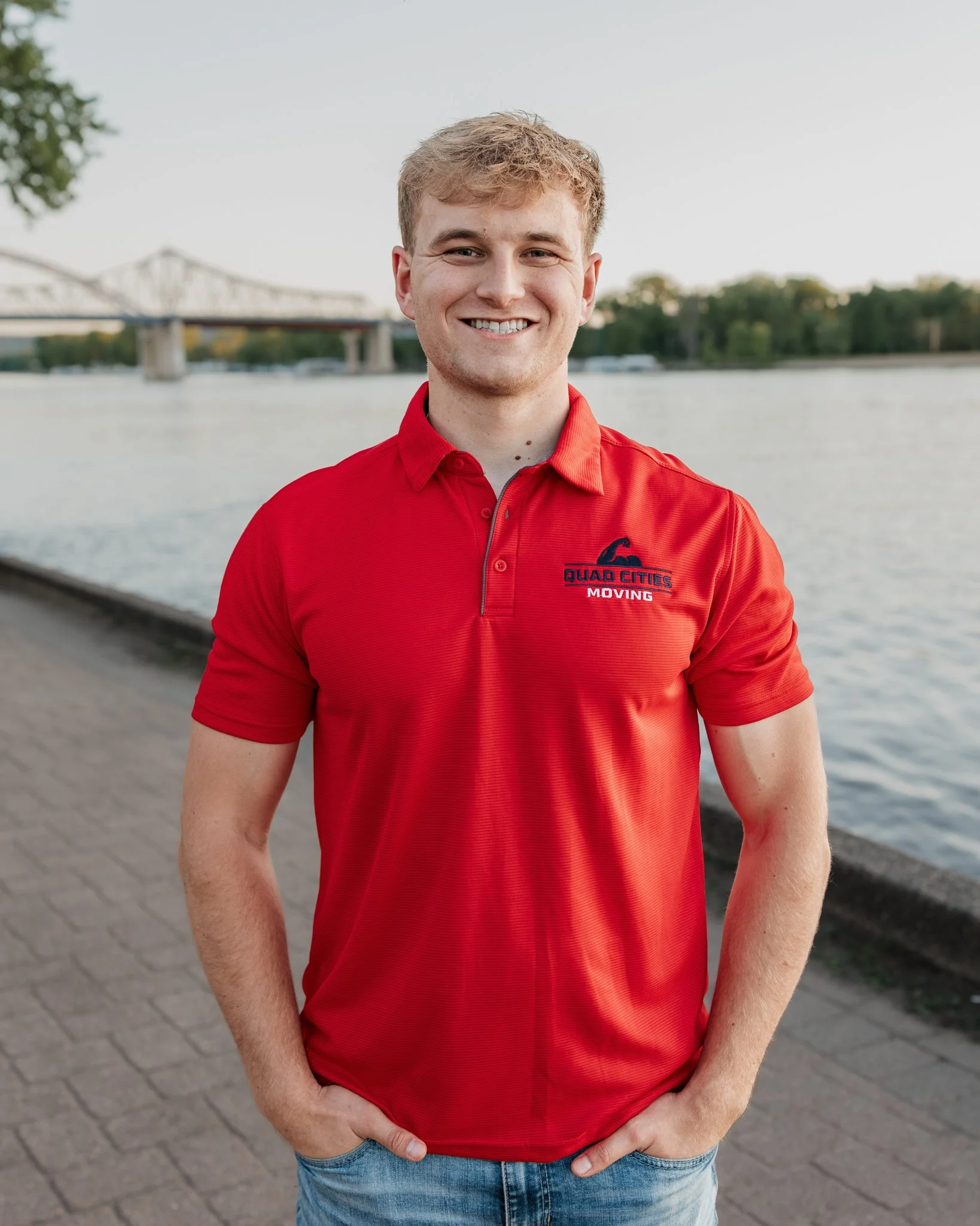 Young man smiling outdoors near a river with a bridge in the background, wearing a red collared shirt with 'Quad Cities Moving' logo.