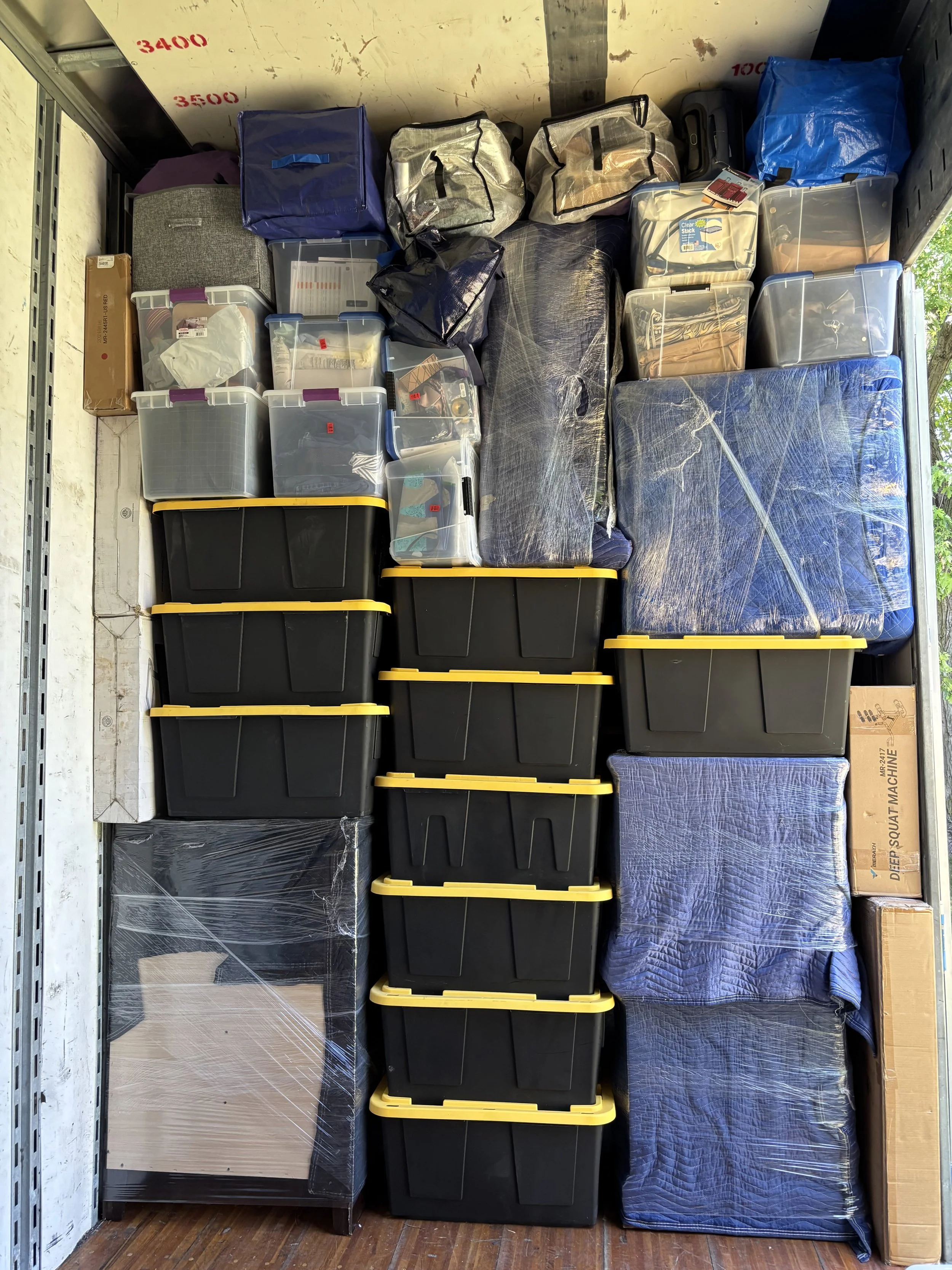 A storage unit with black plastic bins with yellow lids, cardboard boxes, and various wrapped and unwrapped items.