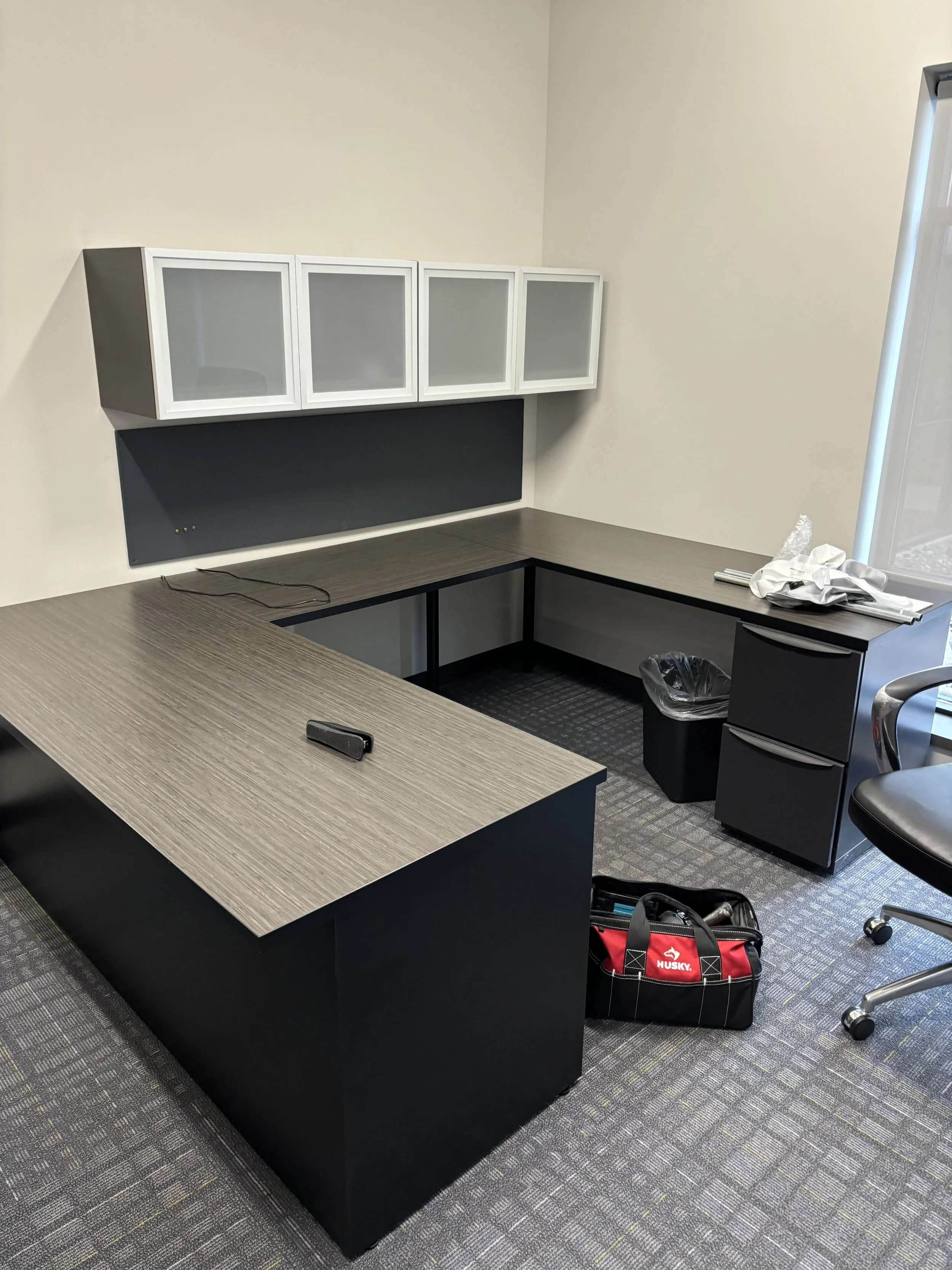 Empty office space with an L-shaped desk, black and gray color scheme, a white wall, overhead cabinets with frosted glass doors, a trash can, tools, and a Husky brand tool bag on the carpeted floor.