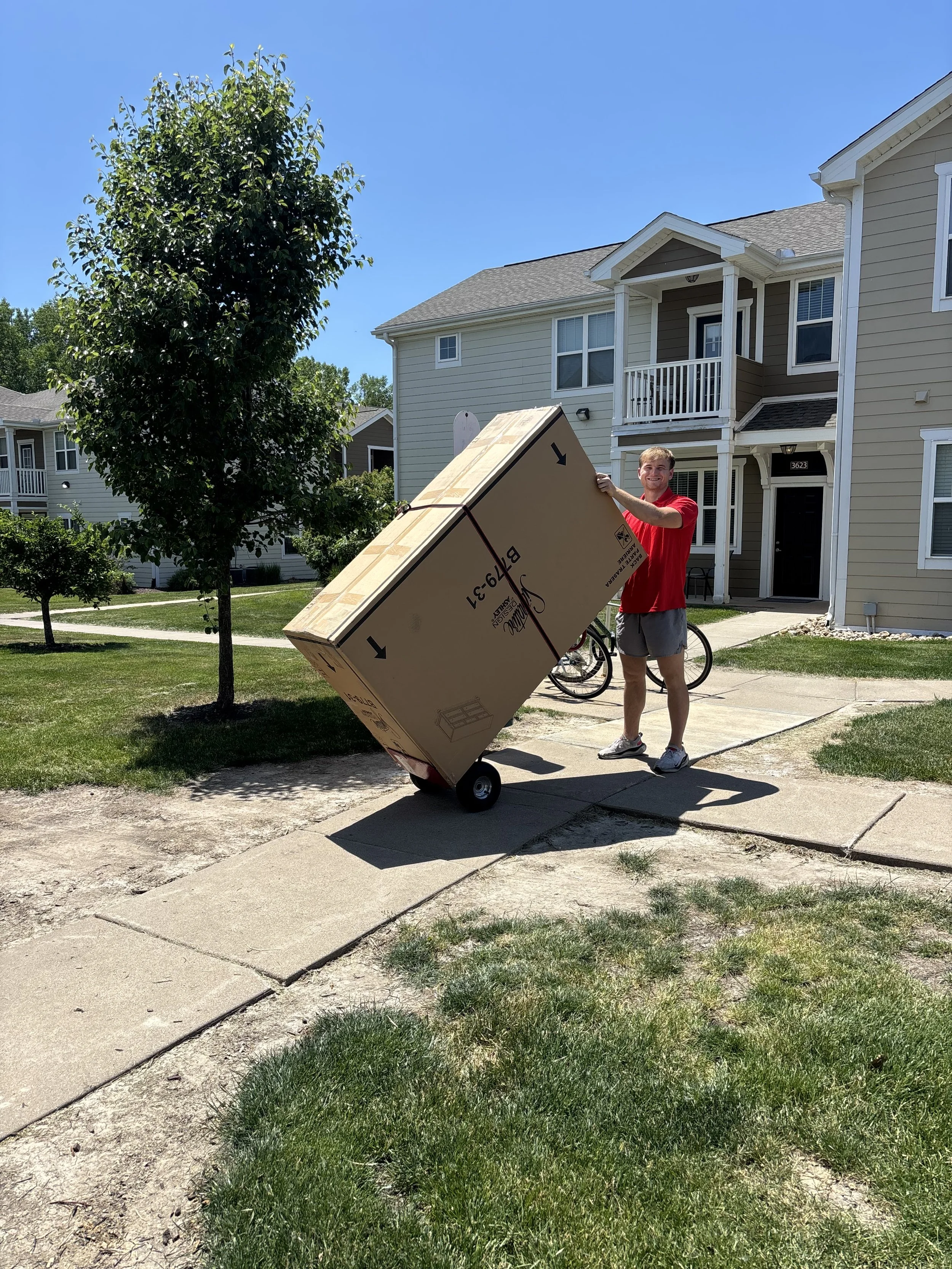 A smiling young man in a red t-shirt and shorts, holding a large cardboard box on a hand truck, standing on a sidewalk in front of a two-story residential building with beige siding, white trim, and a small porch, on a sunny day with a clear blue sky.