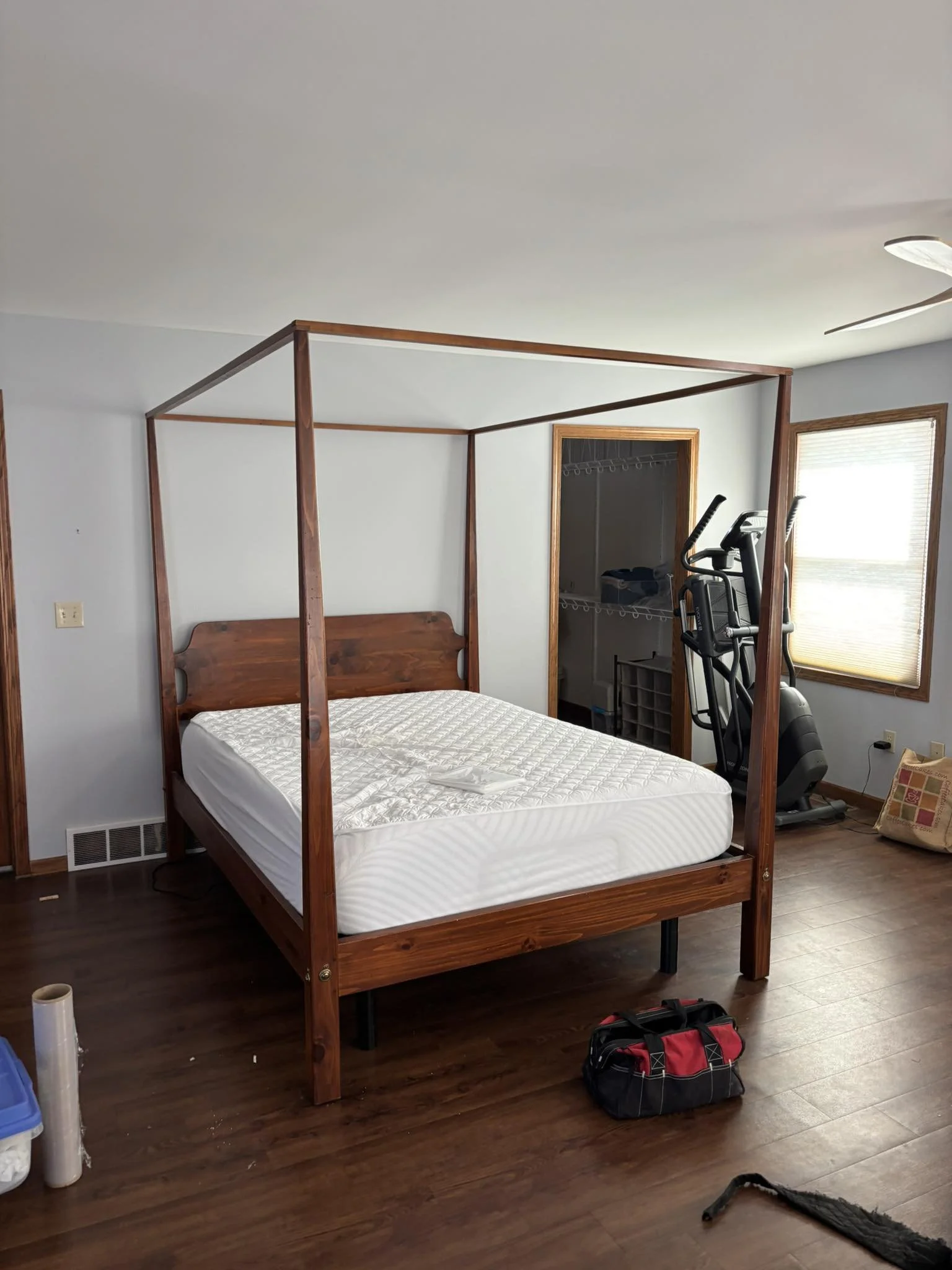 A bedroom with a wooden bed frame without a mattress, a treadmill near a window, and a closet with shelves inside. There is a bag and some tools or objects on the floor, with natural light coming through the window.
