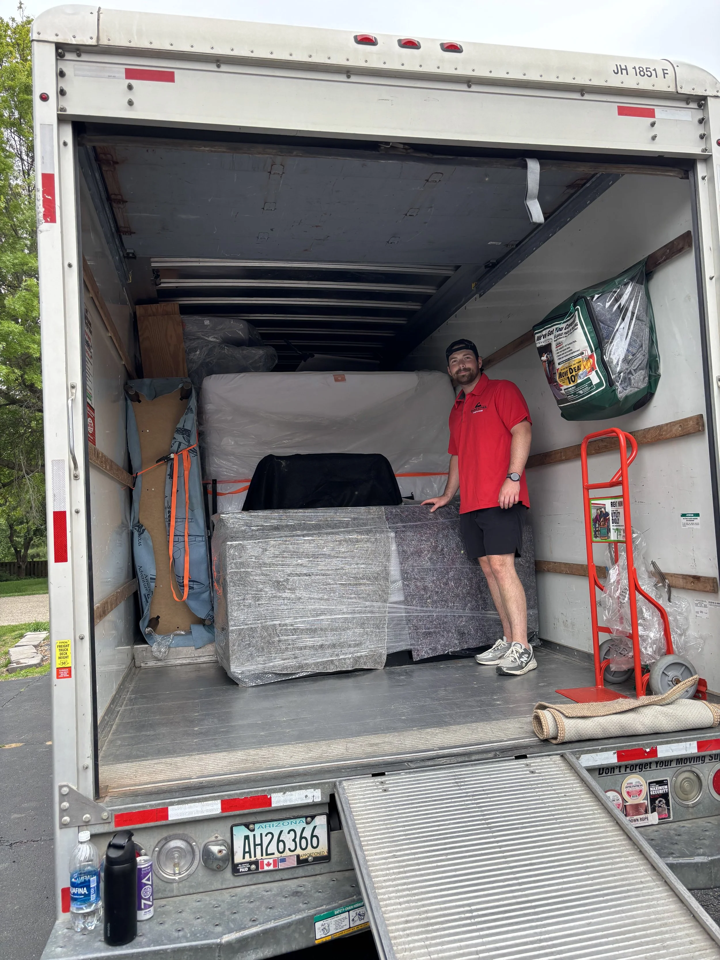 A man standing inside an empty moving truck with various moving supplies around. The man is wearing a red shirt and black shorts, smiling at the camera. Moving supplies include a dolly, plastic bags, bottled water, and other packing items.