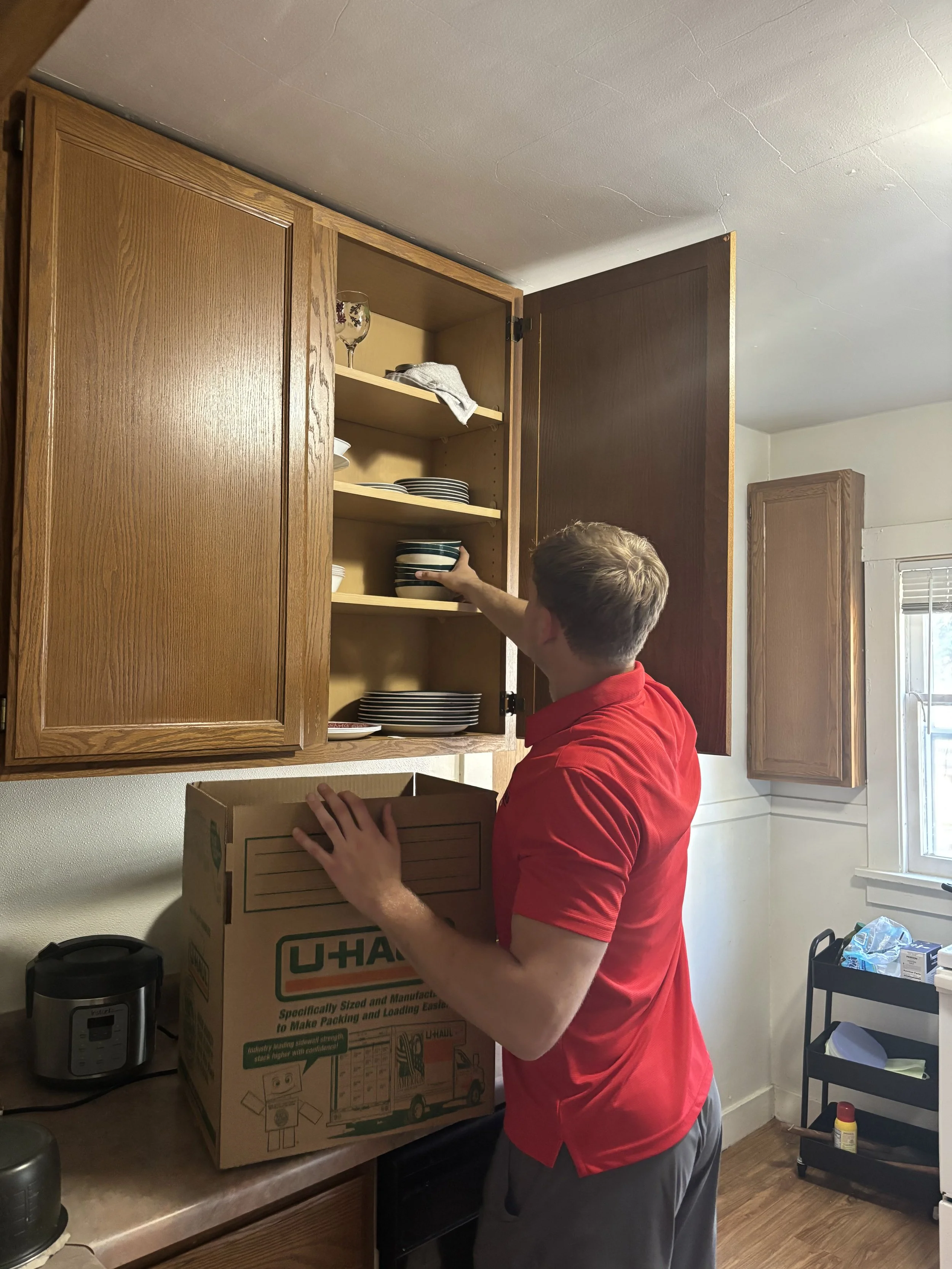 A man in a red shirt is packing a moving box in a kitchen. The kitchen has wooden cabinets, a window, and a small black trolley with cleaning supplies. The man is placing a bowl on a shelf inside the cabinet.