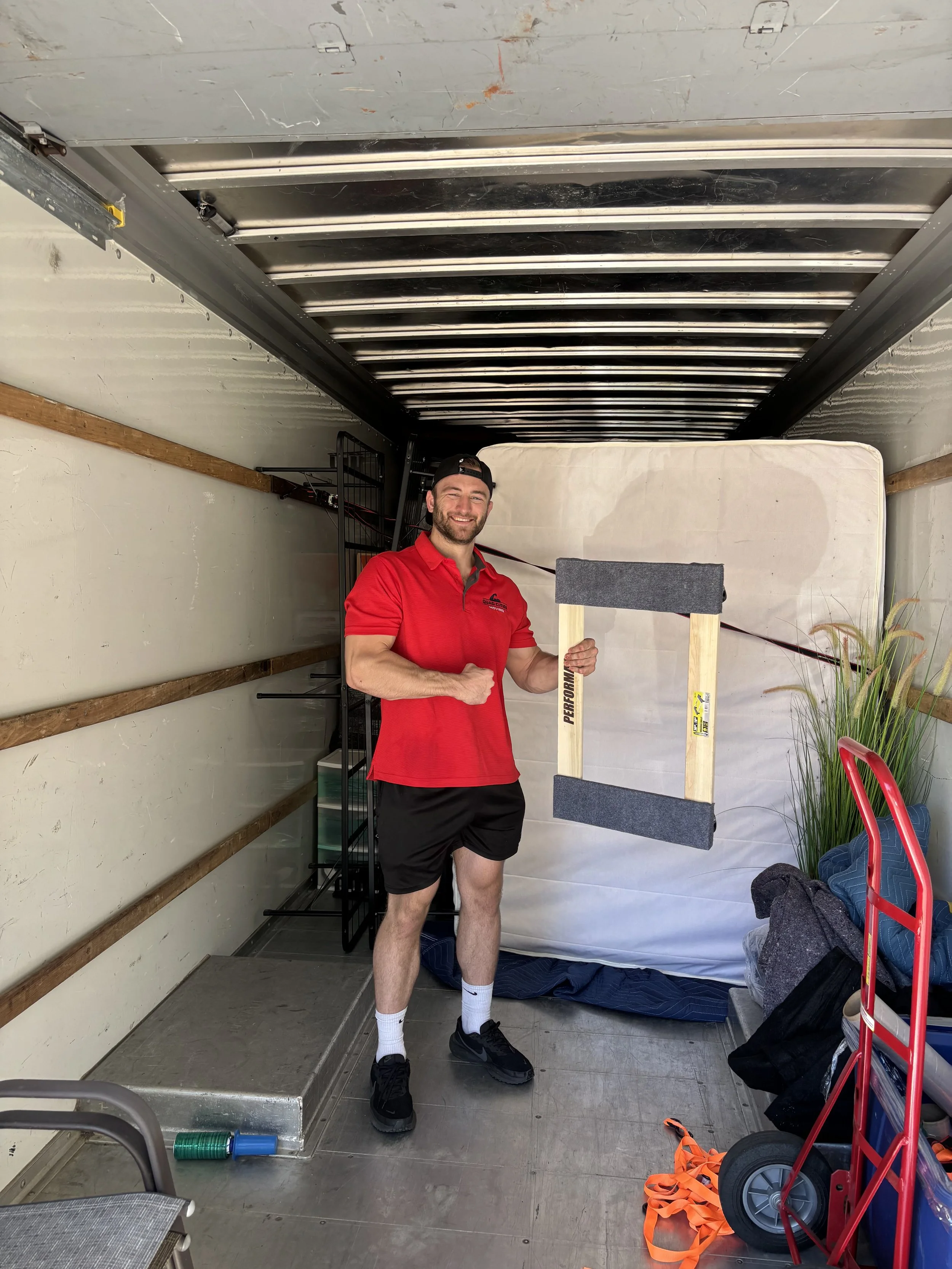 A man with a beard, wearing a black cap backwards, a red polo shirt, black shorts, and athletic shoes, is smiling and holding a wooden sled in a truck's cargo area. The cargo area has a mattress, some plants, bags, and equipment, with a metal ramp at the back.