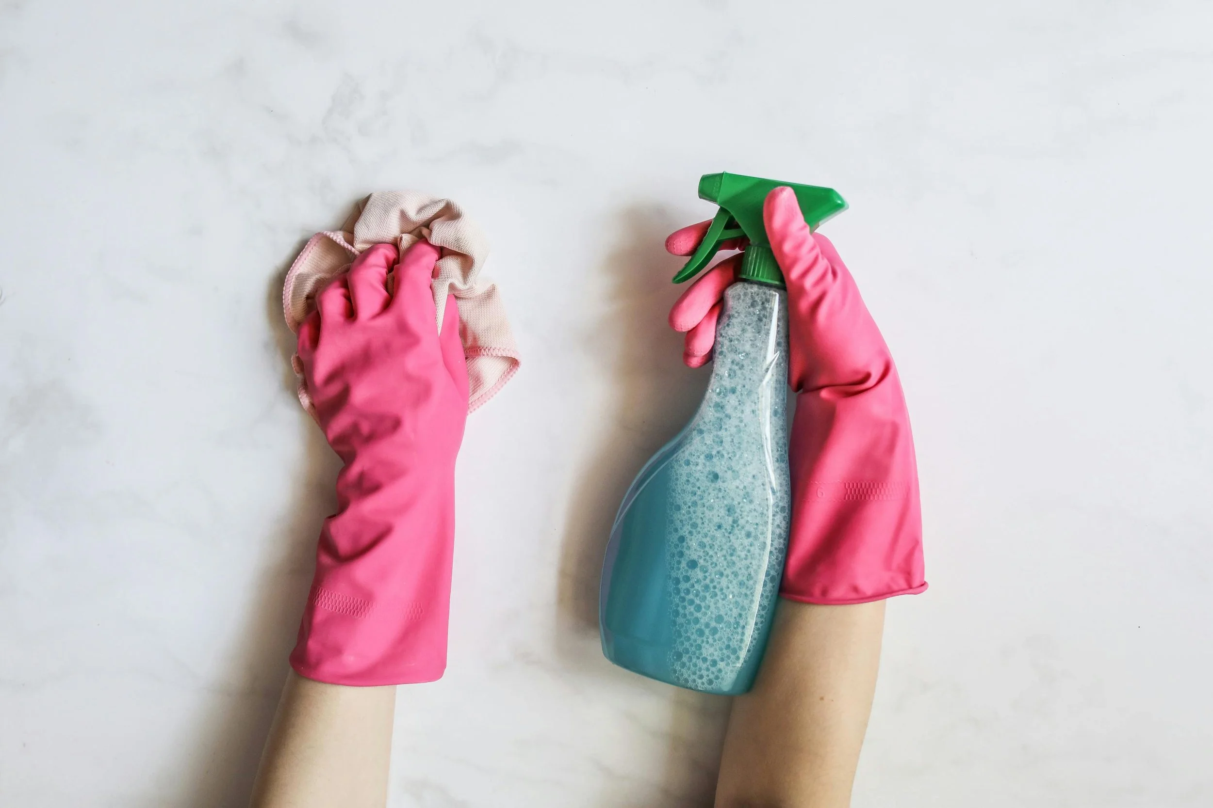 Person wearing pink rubber gloves cleaning a light-colored surface with a spray bottle of cleaning solution and a cloth.