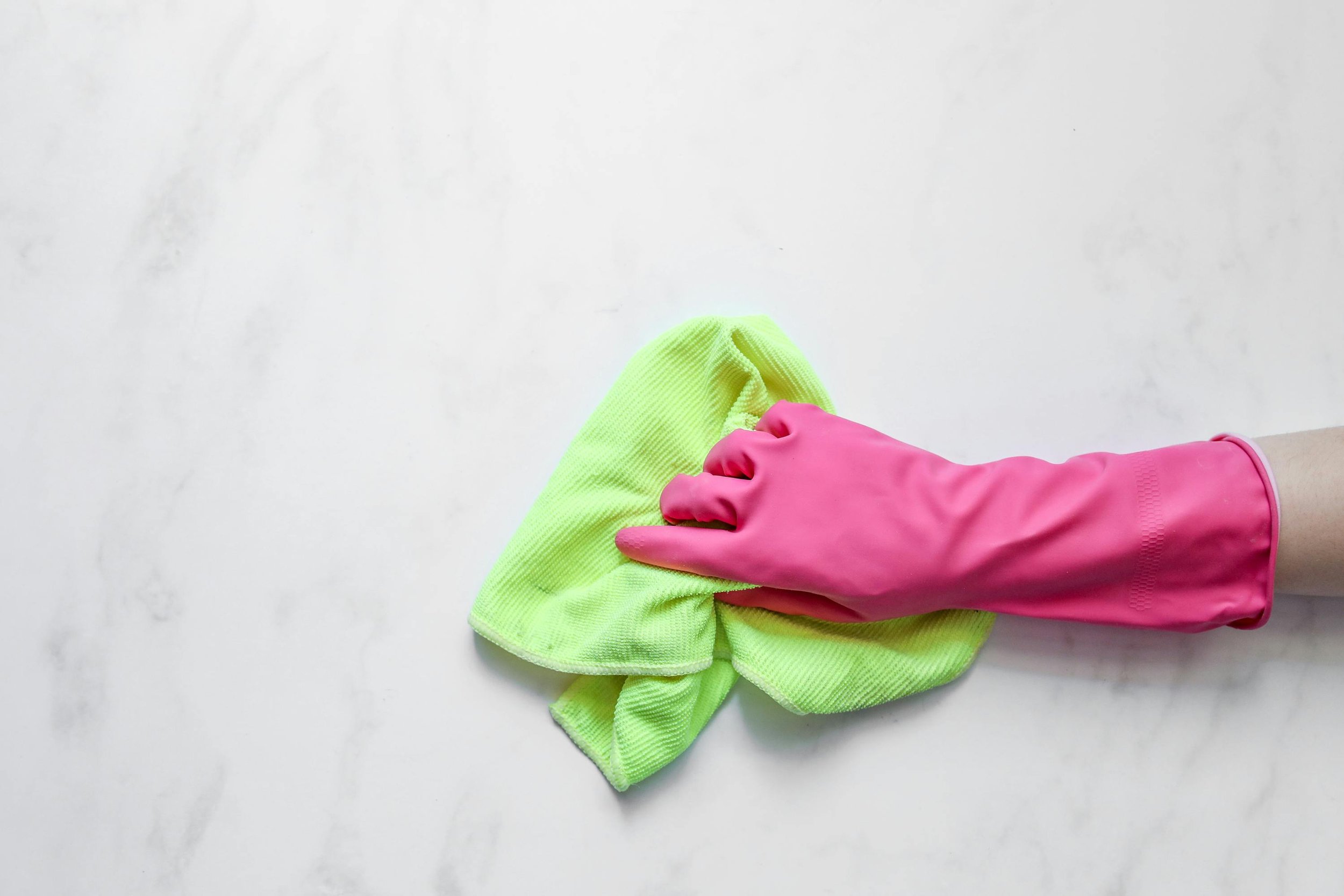 A hand wearing a pink rubber glove is wiping a white surface with a yellow cloth.