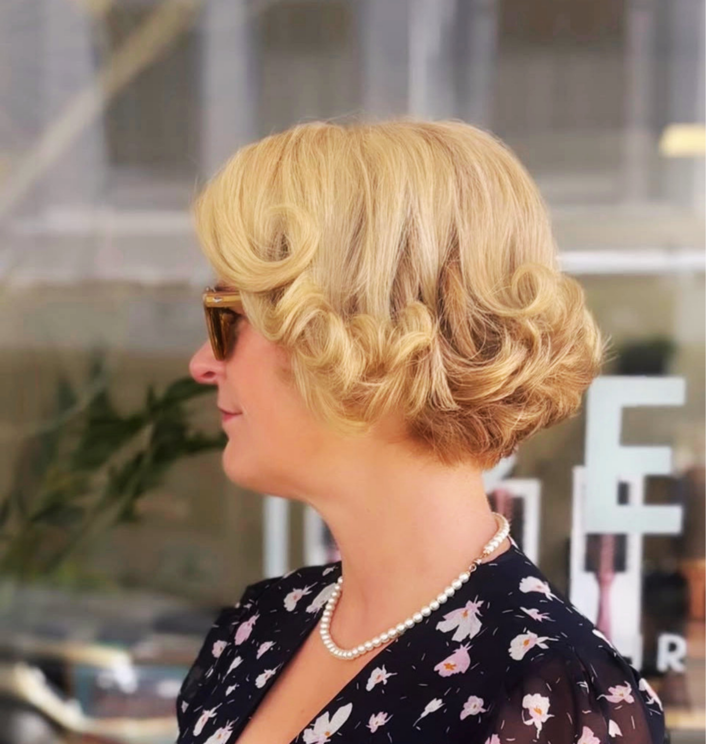 A woman with blonde hair styled in vintage curls, wearing sunglasses, a pearl necklace, and a floral dress.