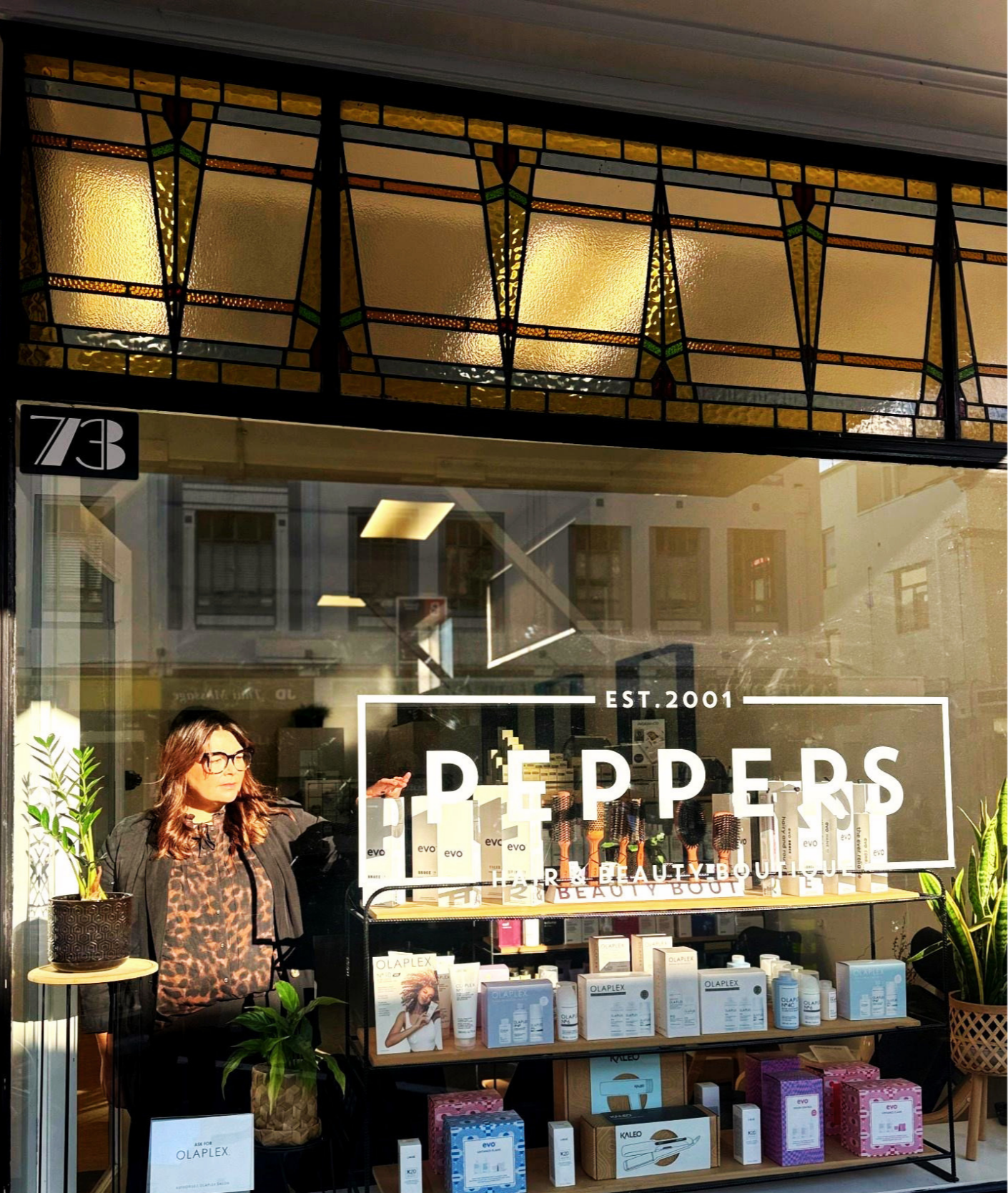Storefront window display for 'Pepper's Hair & Beauty Boutique' established in 2001, featuring hair and beauty products like Olaplex and Evo, with potted plants and a woman inside looking out.