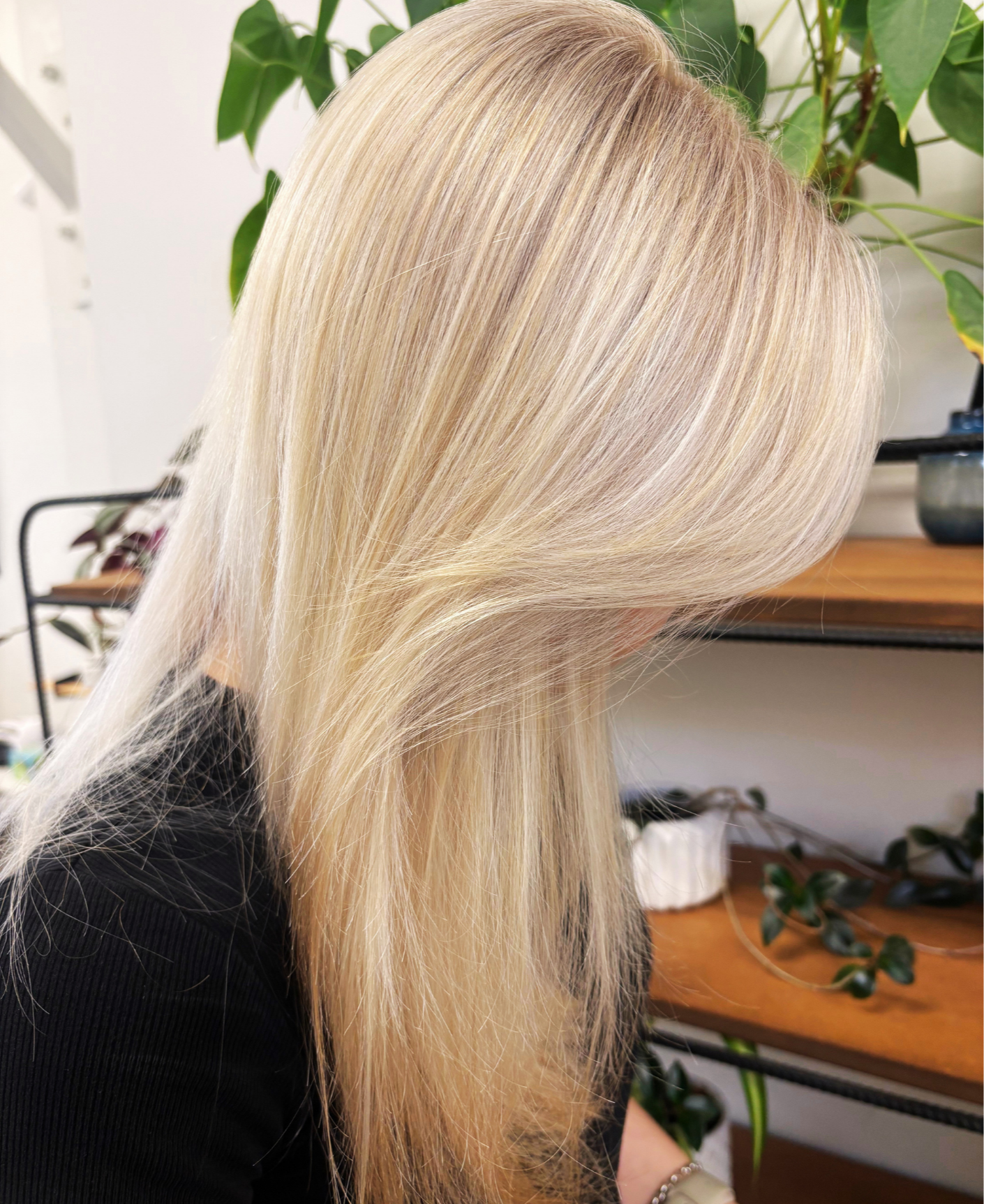 Close-up of a woman with long, straight, blonde hair in a hair salon, with green plants in the background.
